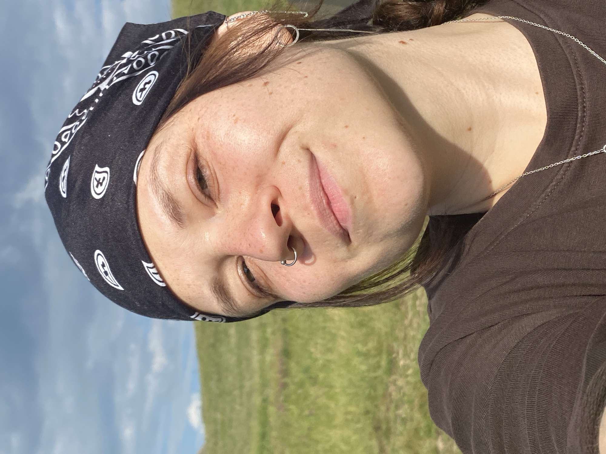 A close-up, outdoor, selfie-style photograph features a young person wearing a black bandana with a white paisley pattern and a brown, ribbed t-shirt. They have a silver hoop nose ring and a dangling silver earring in their right ear. Their hair is brown, and a braid is visible. The background consists of a grassy, rolling hillside under a blue sky with scattered clouds. The location is identified as Абзаново, Russia.