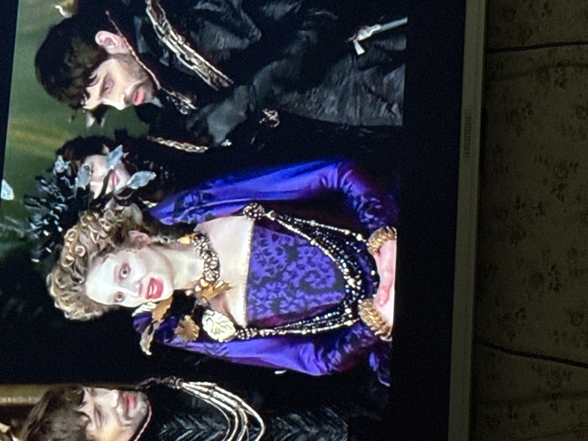 The image shows a television screen displaying a scene with two men and a woman in elaborate costumes. The costumes appear historical, with dark fabrics and gold or silver accents. The woman is wearing a purple and gold dress with ornate jewelry and elaborate hair. The men are in dark clothing with detailed trim. The scene appears to be from a period drama or fantasy film. The television screen is framed by a white border and is situated against a textured wall. The specific location within the city of Mytishchi, Russia, cannot be determined from this image.