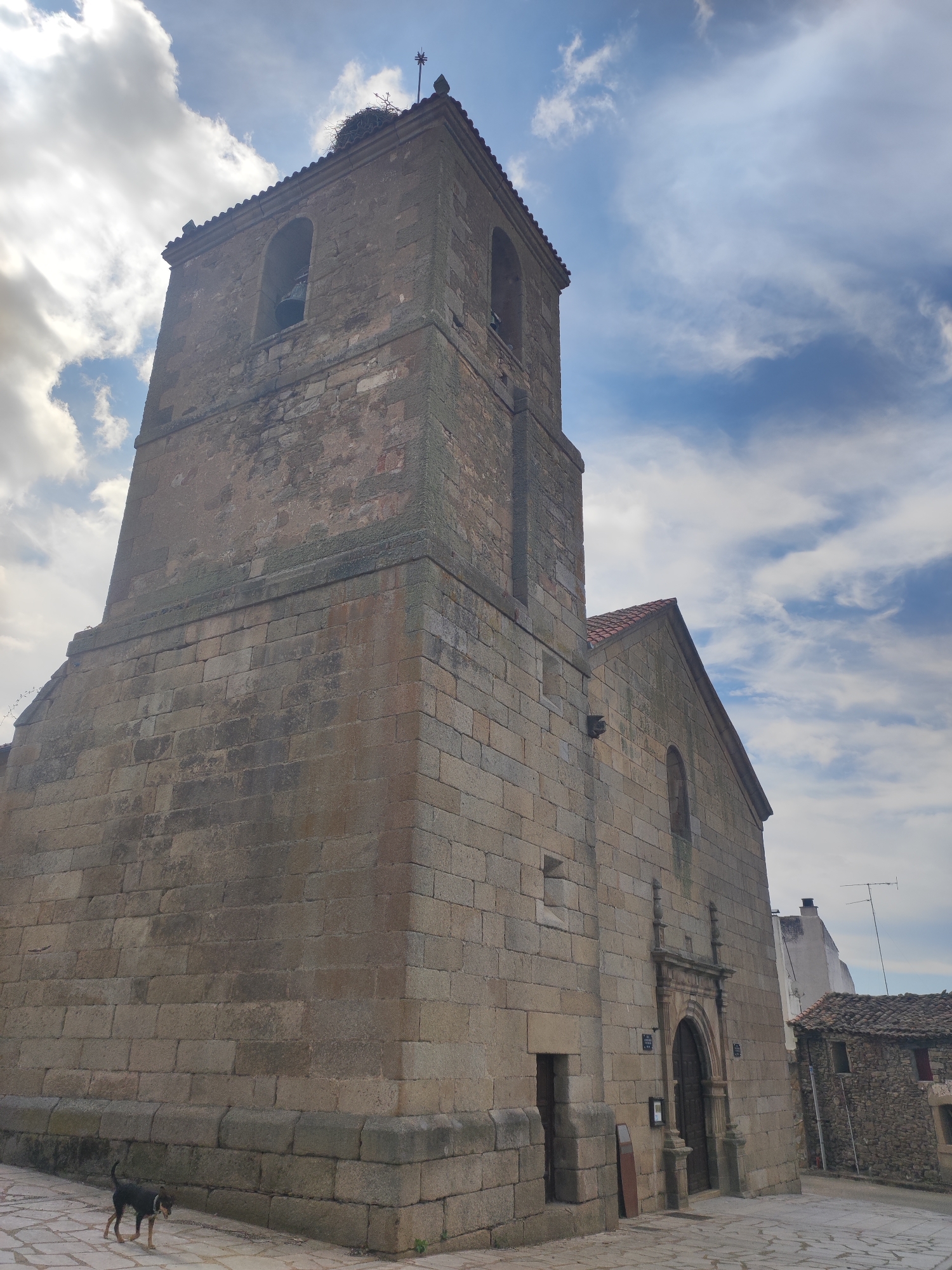 The image shows the exterior of a stone church in Sobradillo, Spain. The church has a tall bell tower with a stork's nest at the top. A bell is visible through an arched opening in the tower. The church building itself is made of large stone blocks and has a pitched roof. A doorway with an ornate stone arch is visible on the side of the church. A small black and tan dog is walking on a paved area in front of the church. The sky is cloudy with patches of blue sky visible. In the background, there are other stone buildings and a white structure with an antenna. The overall impression is of an old, historic building in a rural setting.