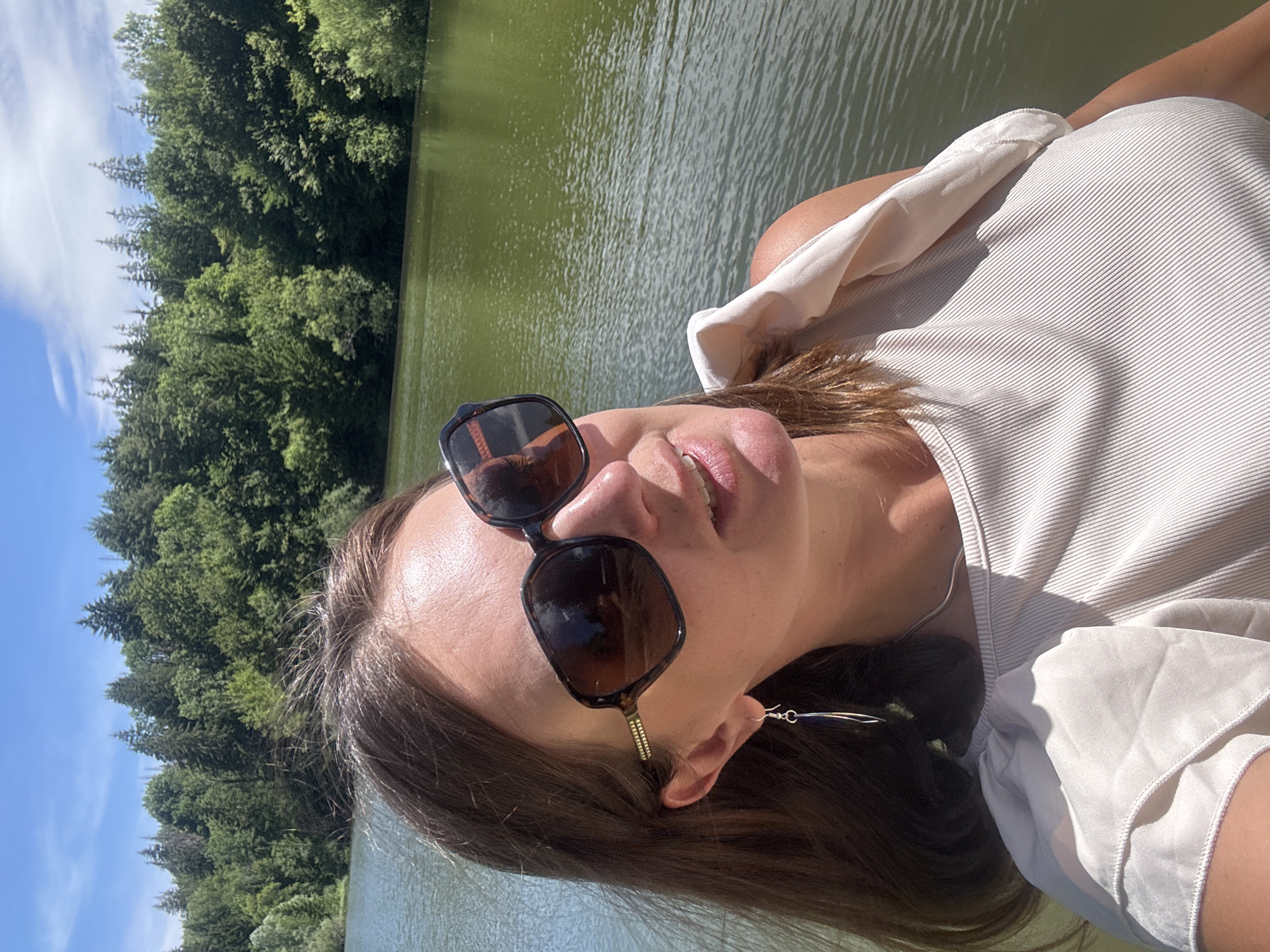 A woman is taking a selfie with a lake and a forest in the background. The woman is wearing sunglasses, a beige top, and silver earrings. The lake is green and calm, and the forest is lush and dense. The sky is blue with a few white clouds. The image appears to have been taken during the daytime on a sunny day. The location is Coteaux du Lizon, France.