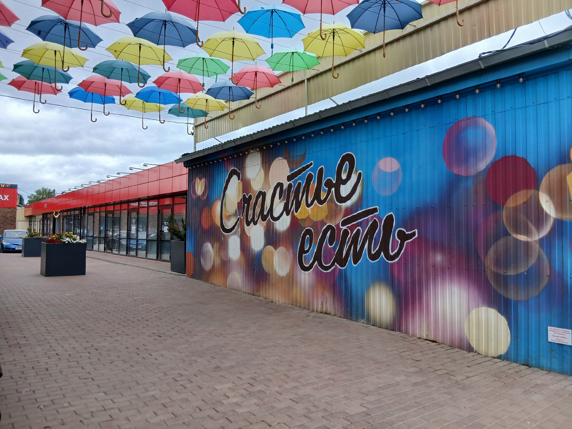 In Osa, Russia, a street scene features an outdoor area with a patterned brick walkway leading towards a building with a red facade. Above the walkway, numerous colorful umbrellas are suspended, creating a decorative canopy against a cloudy sky. To the right, a blue corrugated metal wall is adorned with graffiti depicting blurred circles of light and the Russian phrase "Счастье есть" (Happiness exists). In the background, a blue car is parked near the building's entrance. Planter boxes with foliage are placed along the walkway.