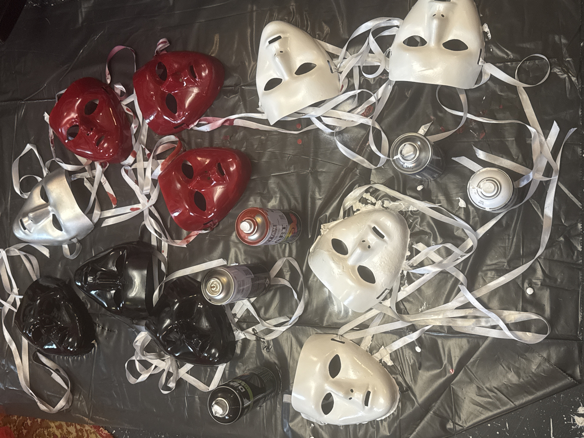 The image depicts a collection of masks and spray paint cans arranged on a black plastic sheet. The masks are of various colors, including black, silver, red, and white. Some of the masks have white ribbons attached to them, which are also scattered across the sheet. There are three spray paint cans visible: one black and white, one silver, and one red and white. The overall scene suggests a crafting or art project involving the decoration of masks. There is no indication of the specific location being Komsomolsk-on-Amur, Russia, nor any information about people, actions, or interactions.