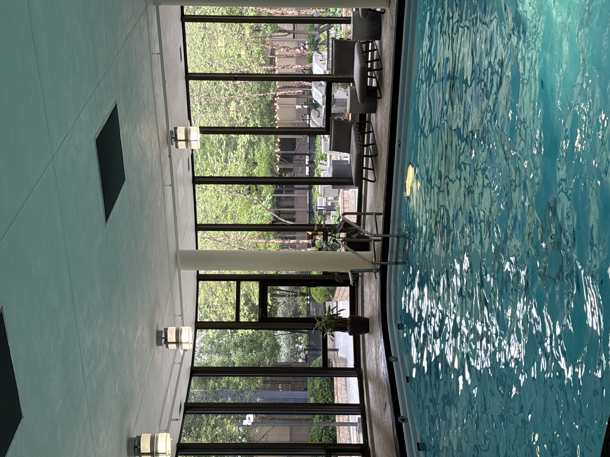 This image depicts an indoor swimming pool area located in the city of Chicago, United States of America. The pool itself is filled with turquoise water, with visible ripples and reflections on its surface. The pool is surrounded by a dark border and a light-colored tiled deck.
Large windows line the perimeter of the pool area, offering views of an outdoor courtyard or patio. Through the windows, one can see trees with lush green foliage, outdoor seating arrangements including chairs and tables, and what appear to be outdoor grills. The architecture visible through the windows suggests an urban setting, possibly a residential complex or hotel.
Inside the pool area, several lounge chairs with gray cushions are positioned near the windows. There are also cylindrical planters with greenery, one housing a large potted plant near the pool's edge. The ceiling is light-colored and features recessed lighting fixtures, some of which are illuminated. A metal ladder provides access to the pool. The overall atmosphere is serene and well-lit, suggesting a recreational space. There are no people visible in the image.