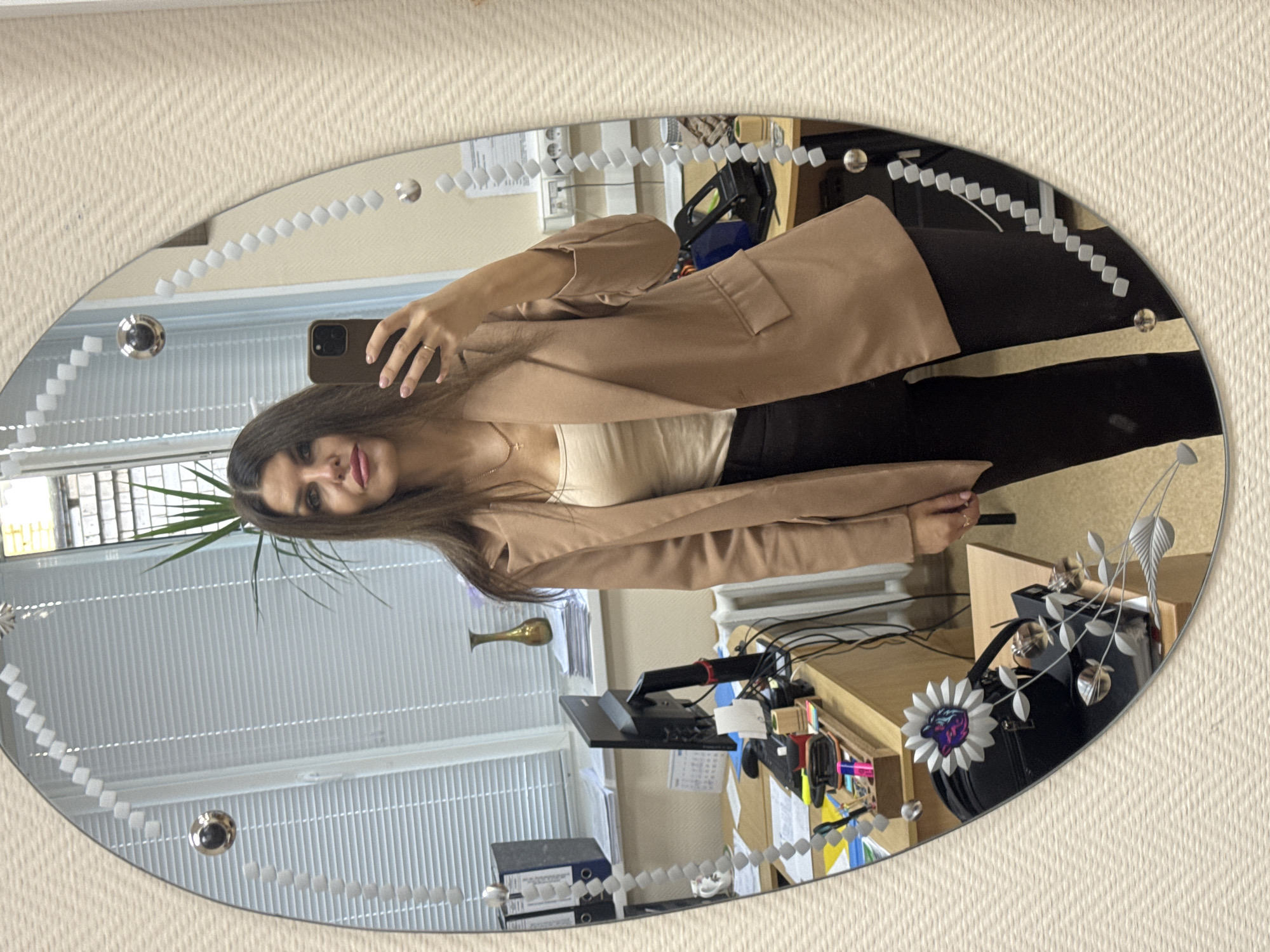 A young woman, with dark hair and fair skin, stands in front of a mirror, taking a selfie. She is wearing a tan blazer over a light-colored top and black pants. Her left hand is raised, holding a smartphone. She is wearing a ring on her left ring finger and a necklace. The mirror is oval-shaped and has a decorative border with small, square shapes and circular elements. The reflection in the mirror shows an office environment. There are desks with computers, papers, office supplies, and a plant visible in the background. A radiator is also present. The wall behind the desks appears to be painted a light color, and there are blinds on the windows. The floor is not visible. The overall lighting suggests an indoor setting, likely during the daytime. The location is identified as Chaikovsky, Russia.