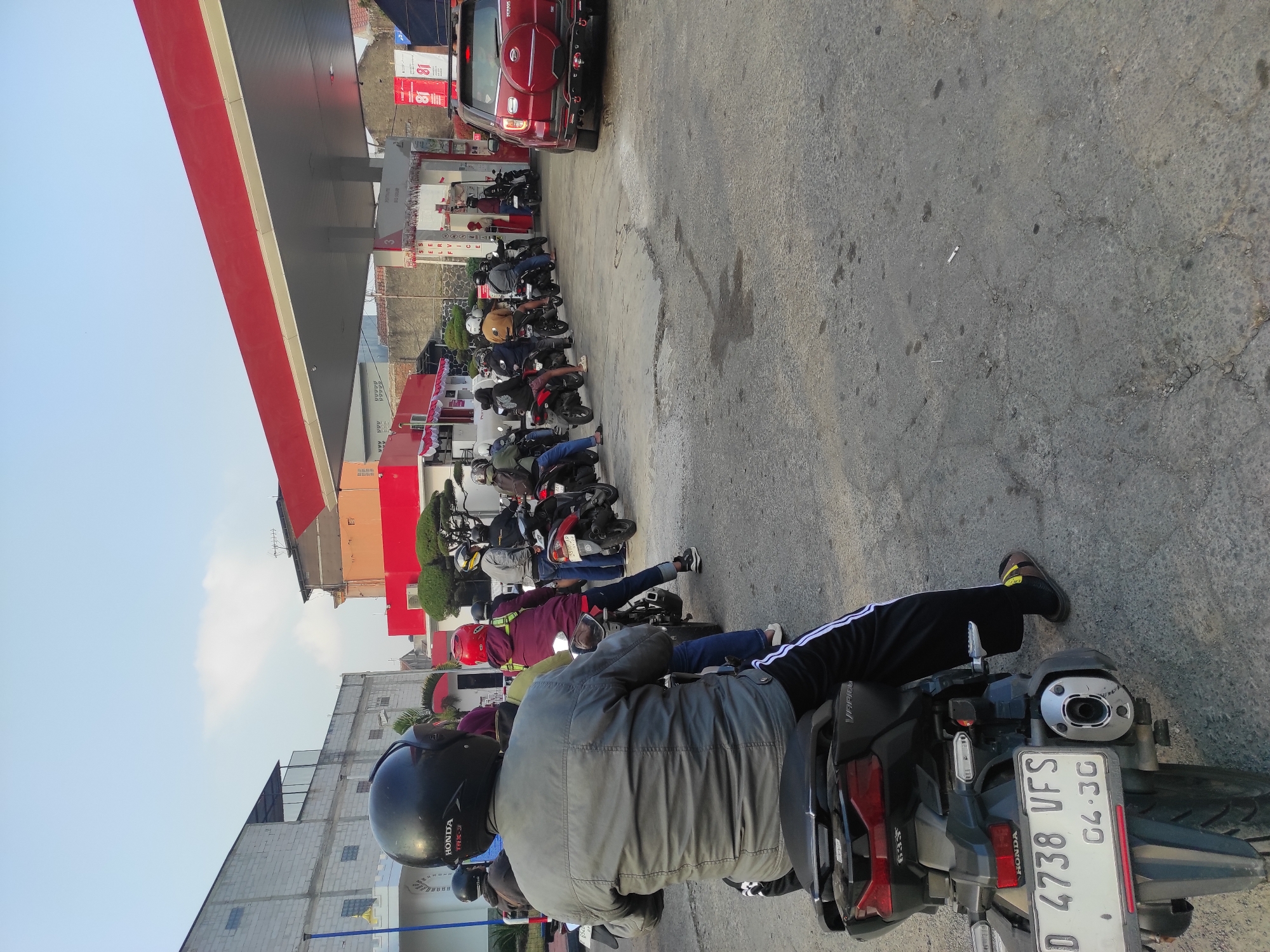The image depicts a line of motorcycles and a red SUV waiting at a Pertamina gas station in Sumedang, Indonesia. The foreground is dominated by the rear of a black motorcycle, ridden by a person wearing a grey jacket and black pants with white stripes. The motorcycle has a white license plate that reads "D 4738 VFS" and "04-30". In the background, a row of motorcycles and their riders are queuing. A red SUV is parked to the right, also waiting. The gas station has red and white signage, and a canopy covers the pumps. The sky is clear blue, and several buildings are visible in the background, including a light-colored building with a white base and a red-roofed structure. The ground is paved asphalt, showing signs of wear and some dark stains. The scene suggests a typical day at a busy gas station.
