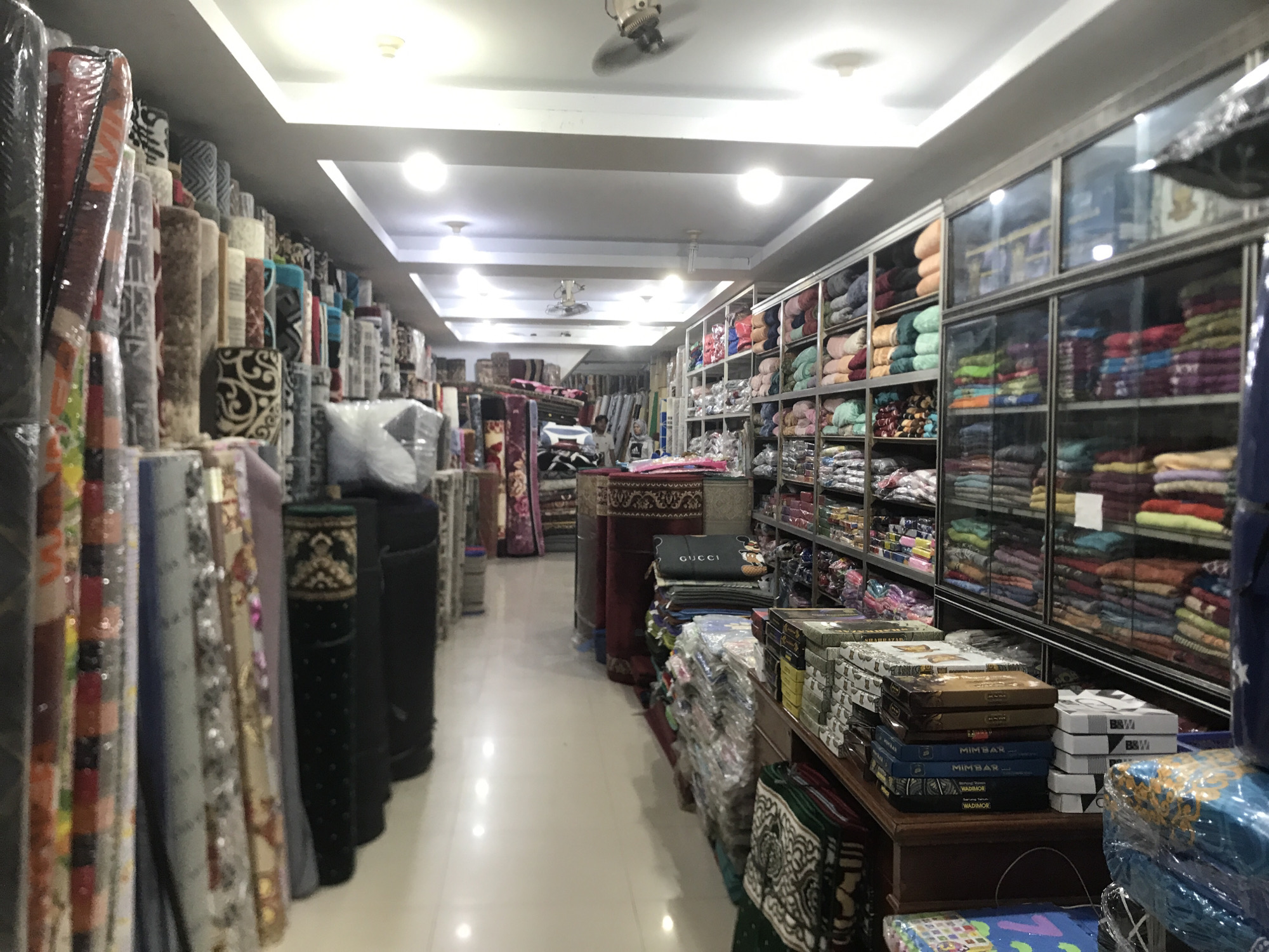 A brightly lit retail space in Banda Aceh, Indonesia, displays textiles. Rolls of carpets stand vertically along the left wall. Along the right, shelves and glass-fronted displays hold stacks of fabrics, while other goods sit on tables and shelves in the aisle. The glossy floor reflects the overhead lighting. A ceiling fan is visible in the center of the image.