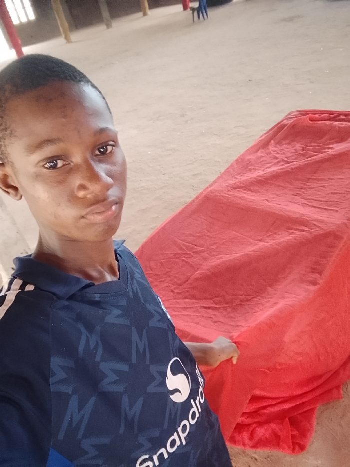 A young male individual, appearing to be of Nigerian ethnicity, is depicted in a medium shot, looking directly at the camera with a neutral expression. He has short dark hair and is wearing a dark blue collared shirt with white stripes on the shoulders, a subtle pattern (resembling 'M' or Sigma symbols), and a white "Snapdragon" logo on the chest. To the subject's right, a large, elongated object is covered by a rumpled, bright red fabric. The subject's right hand is partially visible, resting on this red covering. The ground is a light brown, unpaved dirt surface. In the background, multiple light-colored structural pillars are visible, along with two blue plastic chairs further in the distance, suggesting a large, semi-enclosed, possibly unfinished, or open-air structure. The scene is located in the city of Nteje, Nigeria.