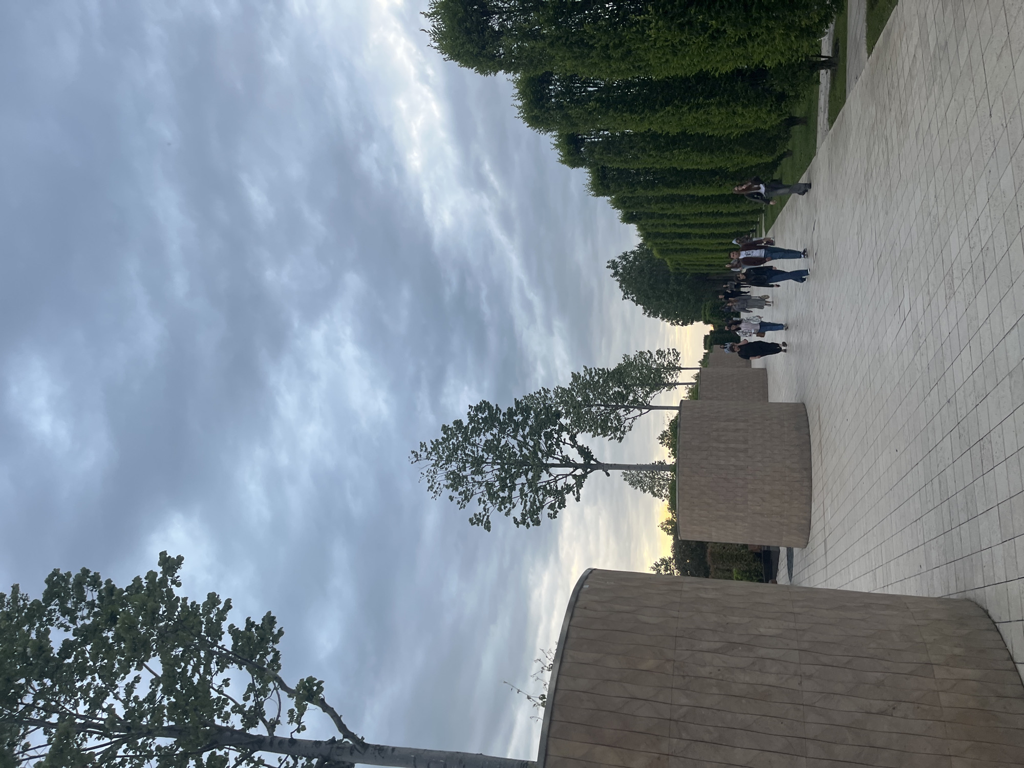 The image depicts a walkway in a park in Krasnodar, Russia. The walkway is paved with light-colored tiles and lined with sculpted shrubbery. A group of people are walking along the path. A modern, low-walled structure made of tan stone with linear carvings is present. The sky is overcast with gray clouds. Trees with sparse leaves are scattered in the park.