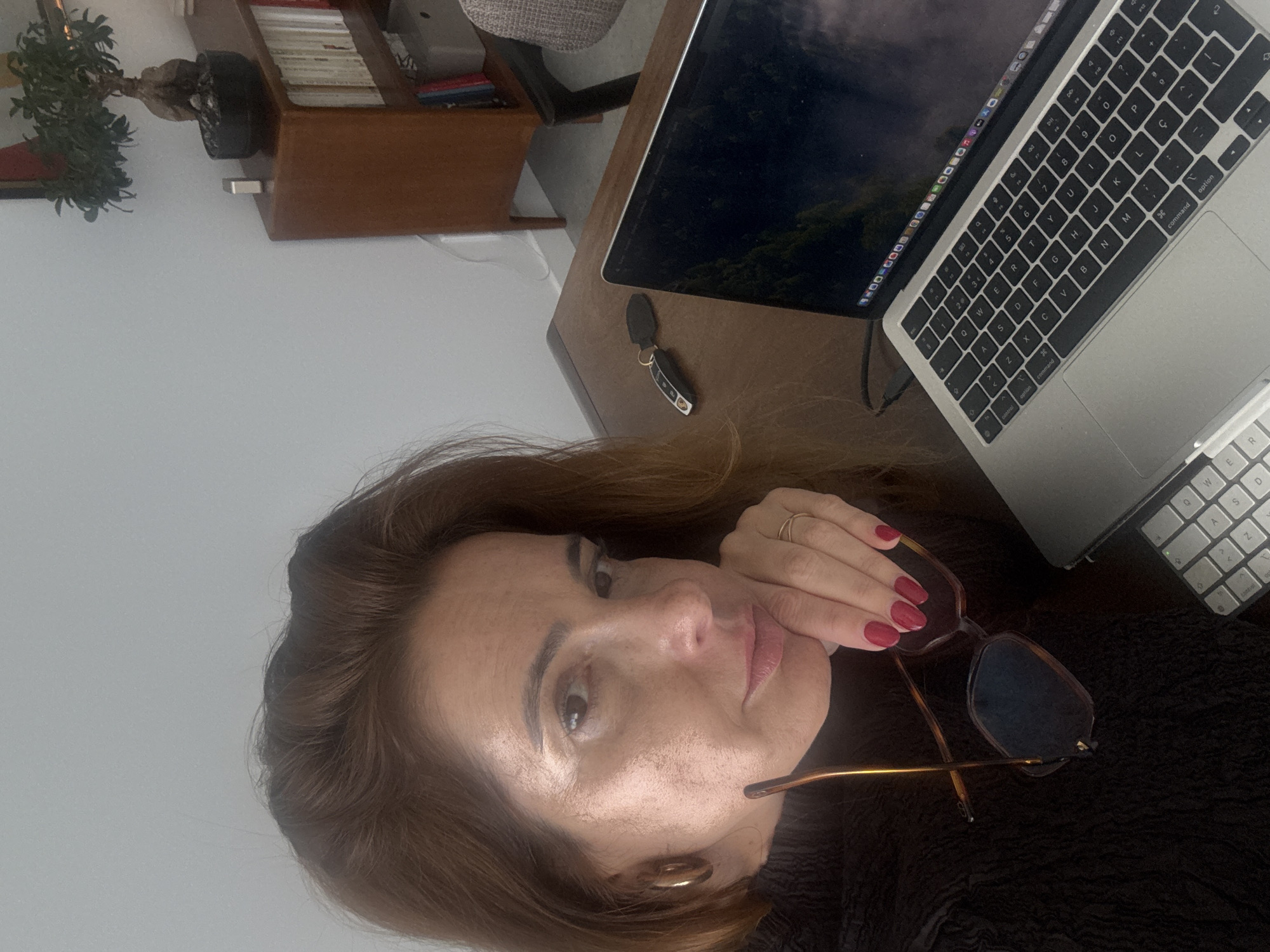 An indoor scene captured in Torres Vedras, Portugal, depicts a female individual with shoulder-length reddish-brown hair and a dark, textured top. She is positioned at a wooden desk, looking directly towards the camera. Her right hand, featuring red nail polish, rests beneath her chin, holding a pair of brown tortoiseshell-patterned eyeglasses with blue-tinted lenses. A gold-colored ring is visible on her right ring finger, and a gold hoop earring is present on her left ear.
On the desk to her right, a silver Apple MacBook laptop is open, displaying a landscape image on its screen. An external white keyboard is partially visible beneath the laptop. A black and silver car key fob is placed on the desk to the left of the laptop. In the background, a wooden bookshelf or cabinet contains multiple books with light-colored spines. A potted bonsai tree and a small, metallic rectangular object rest on top of this cabinet. The walls are light-colored, and a portion of a patterned office chair is visible to the right of the desk.