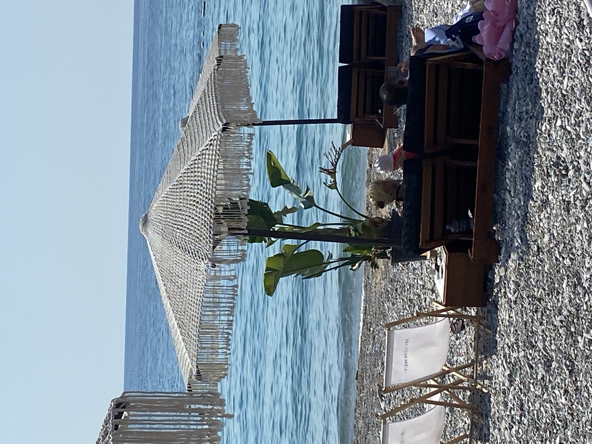 The image depicts a beach scene in Prijevor, Montenegro. The foreground is a pebble beach with several lounge chairs. Two white canvas deck chairs are prominently placed on the left. Behind them, a wooden bench structure holds several individuals relaxing. A large, macrame umbrella with fringe provides shade over this seating area. A potted plant with large green leaves stands near the umbrella pole. In the background, the blue water of the sea meets a clear sky. The sea appears calm, with a few distant buoys visible. The overall atmosphere suggests a relaxed day at the beach.