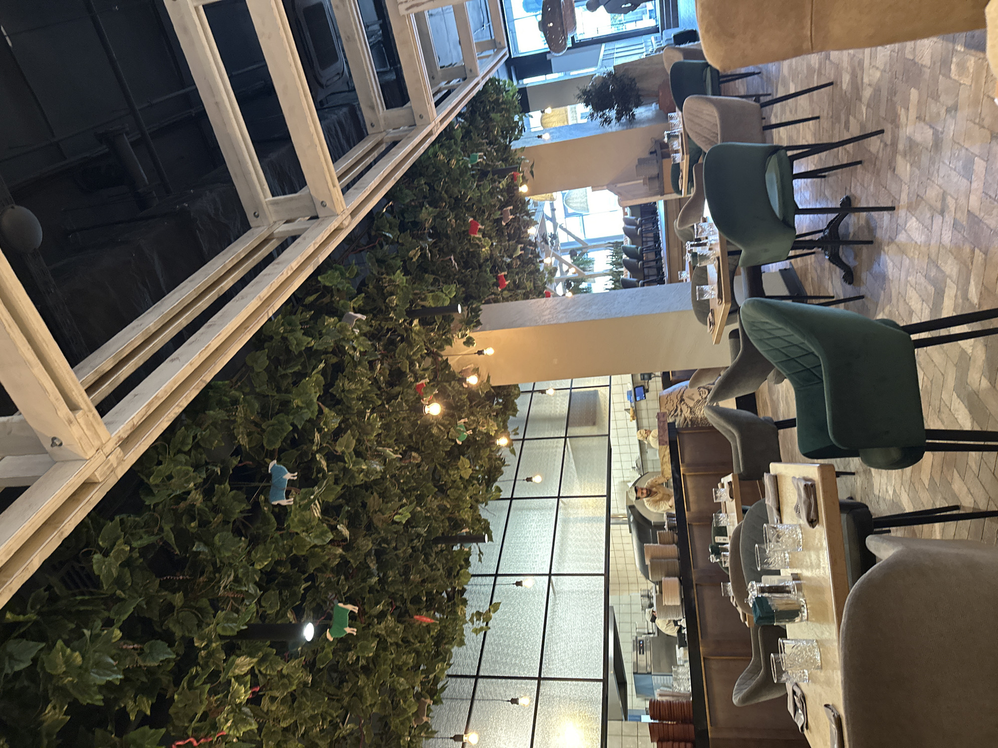 The image shows the interior of a restaurant. A white, open-frame structure is positioned along the left side of the frame, covered with dense green foliage interspersed with small decorative objects, including what appear to be animal figures and small lights. The right side of the frame shows tables and chairs, with place settings on the tables, suggesting a dining area. The chairs are upholstered in shades of green and gray. The floor is tiled in a patterned design. In the background, a window reveals a view of a kitchen area with personnel visible. The location of the image is a restaurant in the city of Izhevsk, Russia. There are no visible people interacting, but the overall impression is of a restaurant interior prepared for service.