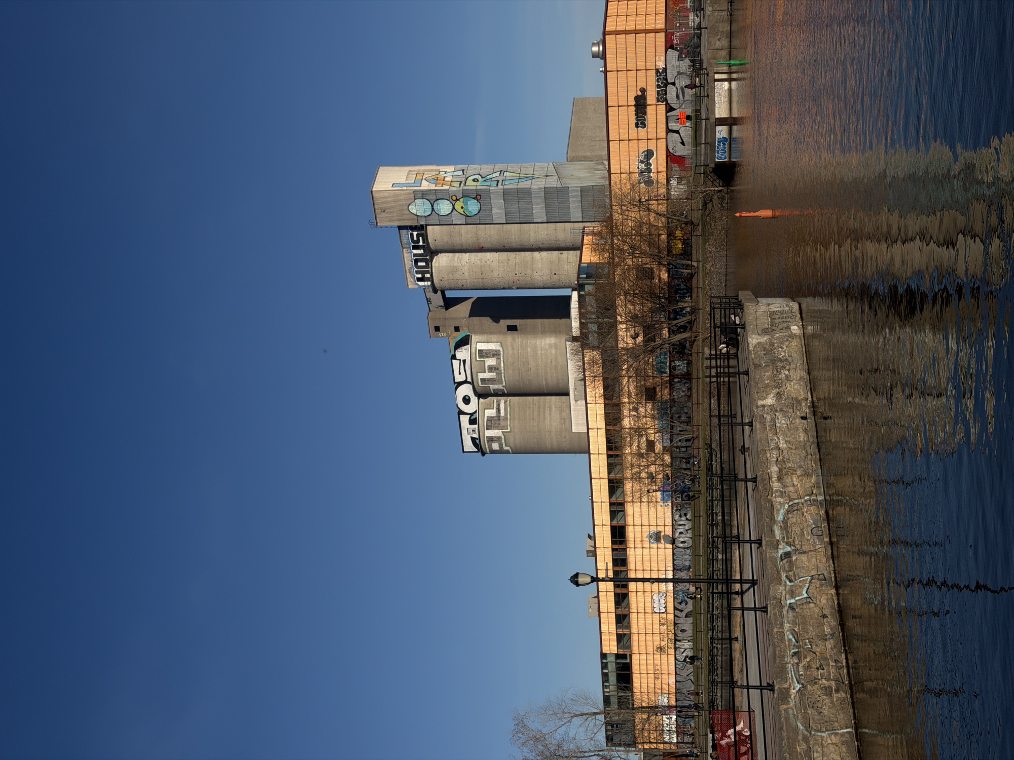 The image shows an industrial complex in Montreal, Canada, reflected in water. The complex consists of cylindrical silos with graffiti and an adjacent building with orange brick walls. A waterfront promenade with railings and trees runs along the water's edge. Graffiti covers the walls and the promenade. The sky is clear and blue.