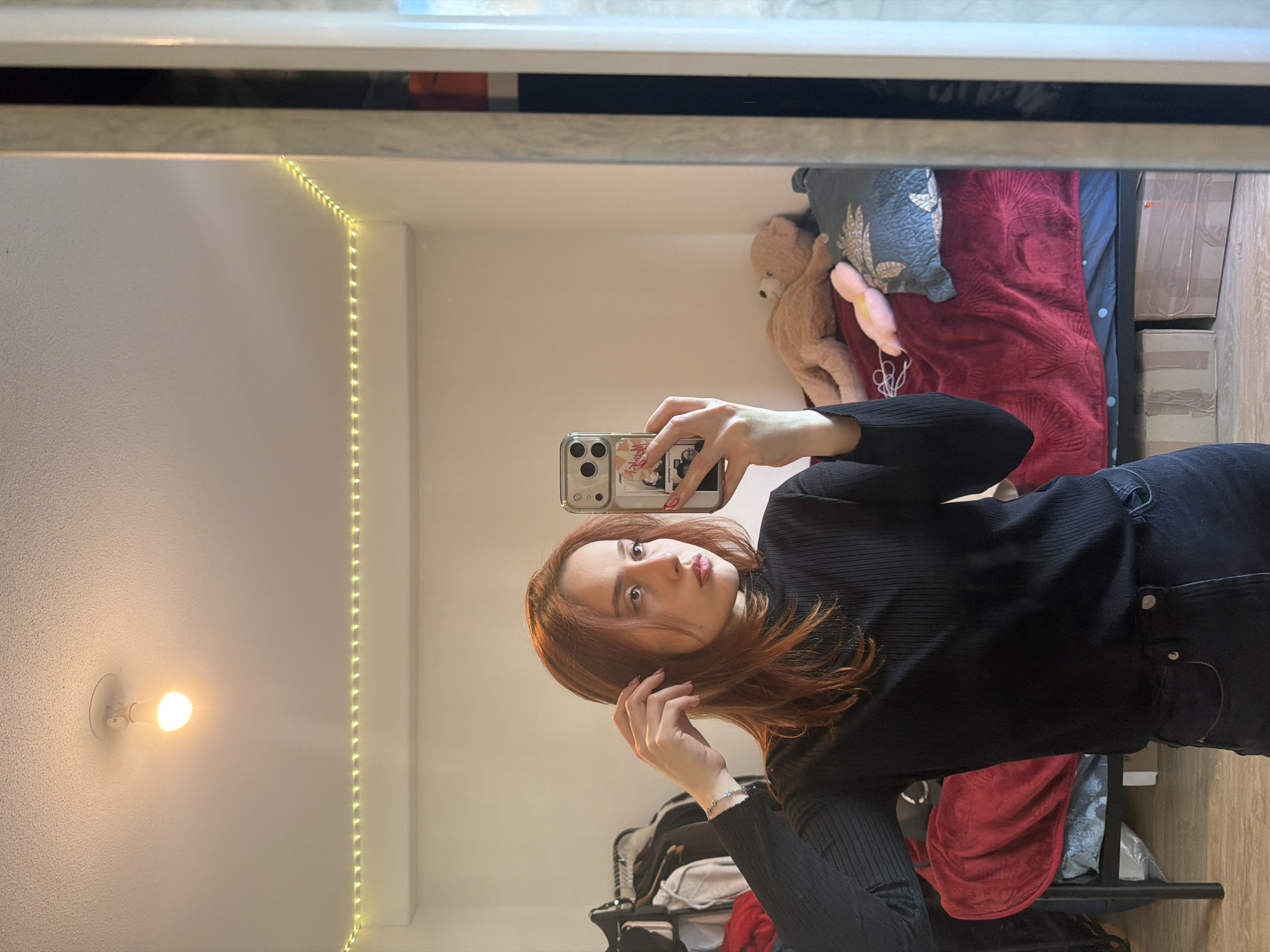 A young female with reddish-brown hair is depicted taking a mirror selfie in a bedroom or dorm room, located in Strasbourg, France. She stands facing her reflection, with her torso and upper legs visible. She wears a black ribbed long-sleeve top and dark jeans. Her left hand is raised, fingers lightly touching her hair near her right ear, revealing a silver-toned bracelet on her wrist. Her right hand holds a silver-colored smartphone featuring a triple camera array, directed towards the mirror. Her fingernails exhibit a light pink polish.
The room's interior, reflected in the mirror, consists of off-white walls and ceiling. Lighting elements include a single, illuminated ceiling-mounted bulb and a string of warm white LED strip lights running along the top of the walls. A bed occupies the background to the right, covered by a deep red blanket over a dark blue fitted sheet with white dots. A light brown teddy bear, a blue-green patterned throw pillow, and a pink, elongated plush object rest on the bed. Two taped cardboard boxes are visible under the bed frame. To the left, a clothing rack partially laden with garments is discernible. The floor is covered with light-colored wood or laminate planks. The mirror itself has a prominent light-colored frame, exhibiting a textured or layered design along its right vertical edge.