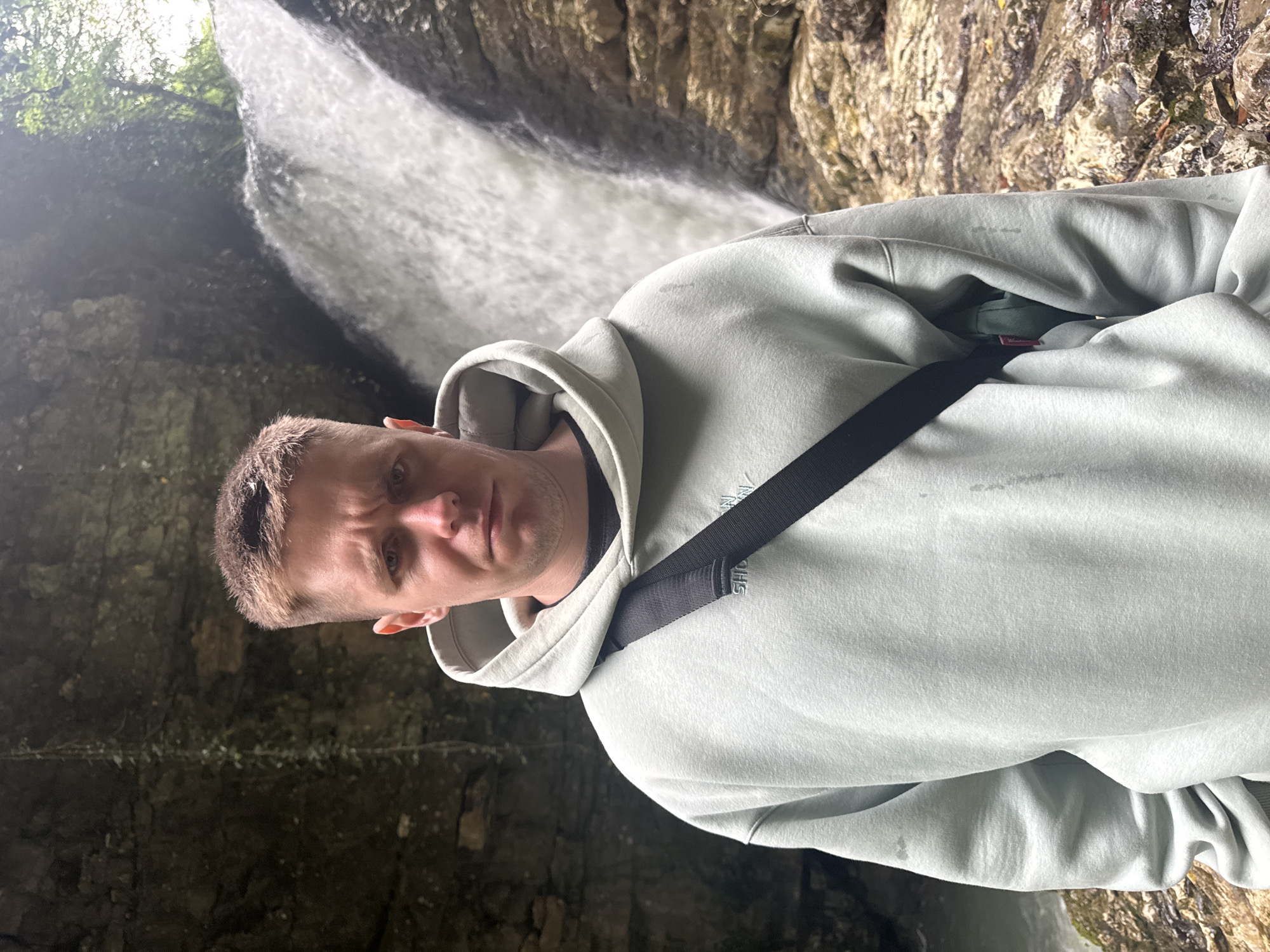 A close-up, medium shot shows a man standing outdoors in a rocky, shaded area with a waterfall in the background. The man is fair-skinned with short, light brown hair and a serious expression. He is wearing a light gray hooded sweatshirt with a black strap across his chest. The location is in Tsabal, Abkhazia, identified by the distinct geological features and waterfall in the background. The environment appears damp and rocky, indicative of a natural canyon or grotto near the waterfall. The photo is taken from a slightly low angle.