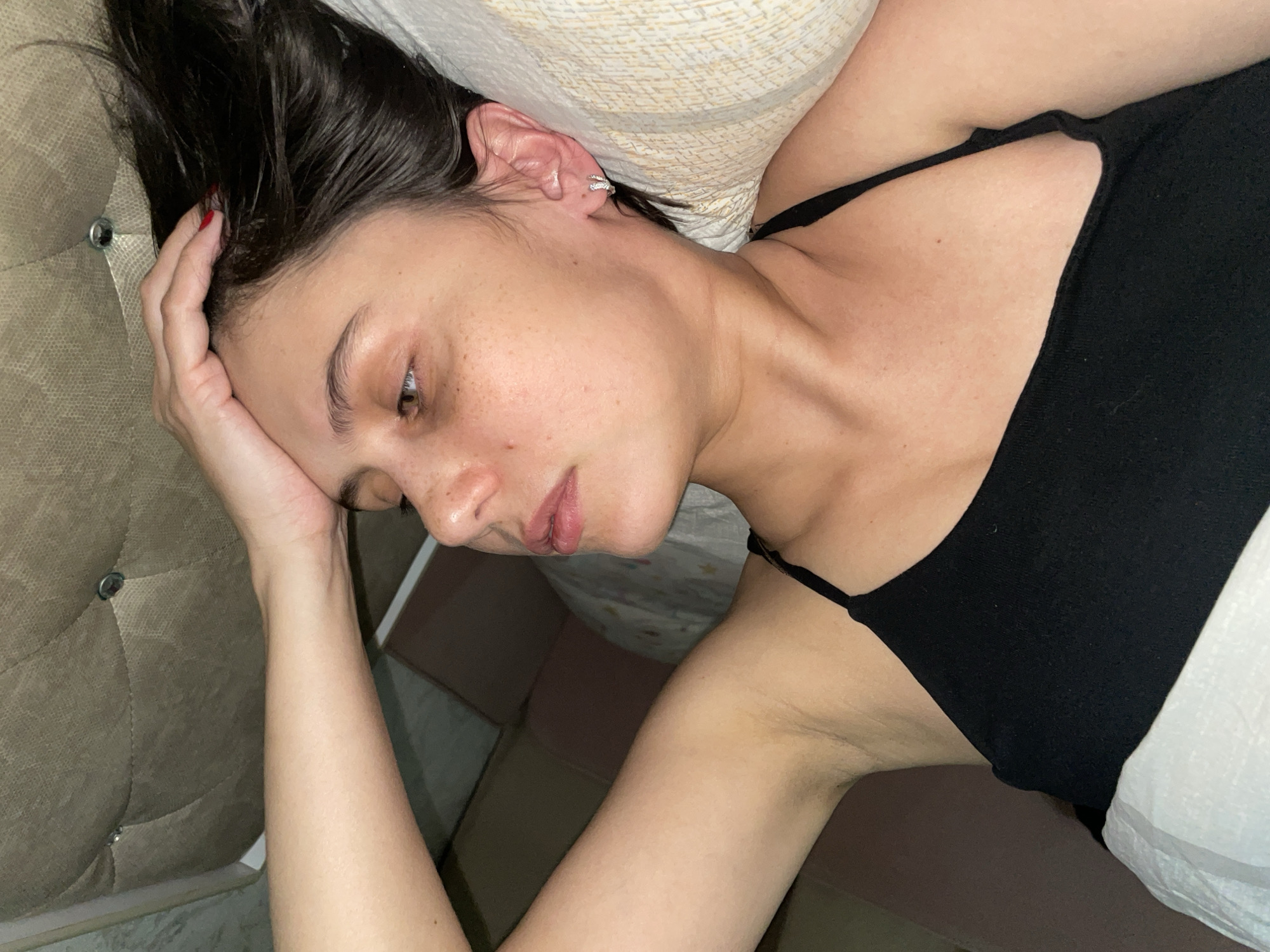 A woman is lying on a bed. She has dark, wet hair and is wearing a black tank top. Her left hand is resting on her forehead. She has fair skin with freckles and a small earring in her left ear. The bed has a tufted headboard and light-colored linens. The background is blurred and shows what appears to be a box and part of a floor. There is no indication of this photo being taken in Izhevsk, Russia.