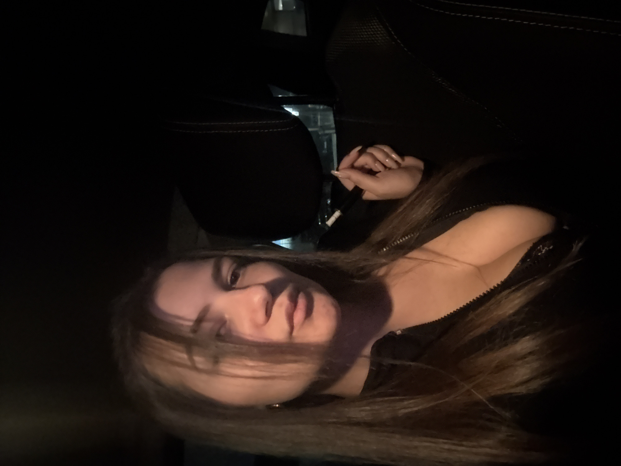 A woman is depicted seated inside a vehicle at night, positioned to the left of the frame, facing the camera directly with a neutral expression. She has long, dark brown hair, with strands partially obscuring her forehead and left eye. She wears a dark, likely black, zippered top or jacket, which is unzipped, revealing a portion of a dark, possibly lace, undergarment. Her right hand, featuring long, almond-shaped fingernails painted a light color, holds a slender object identified as a black and white electronic vaping device or cigarette. To her right and slightly behind her, a dark car seat headrest with distinct white stitching is visible. The background through the vehicle's window is dark and blurred, displaying indistinct lights consistent with an urban environment at night. No specific landmarks are discernible to identify a precise location within Sofia, Bulgaria, beyond an general urban setting.