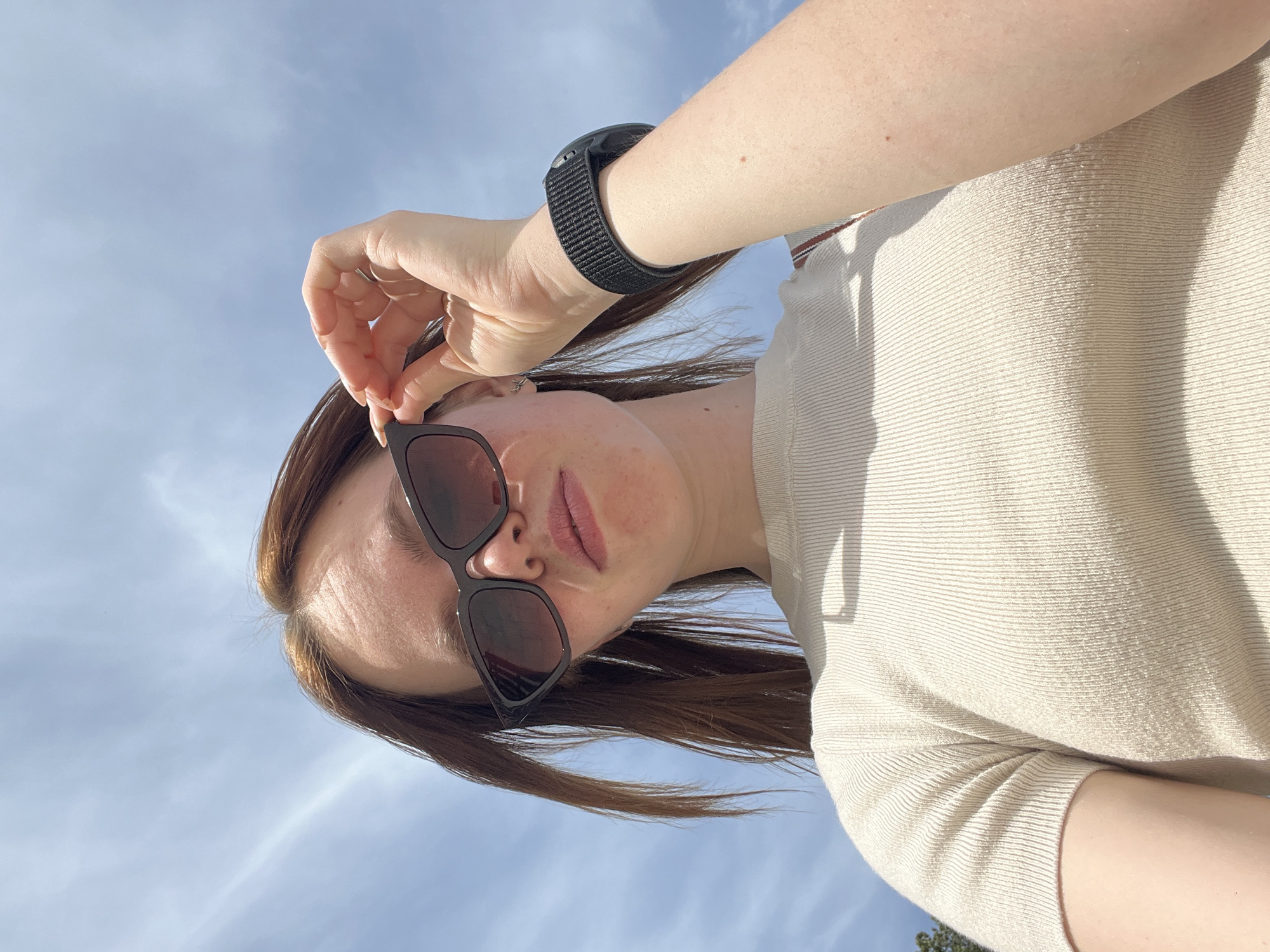 A woman with light skin is centered in the frame. She is wearing dark sunglasses and a light-colored, textured top. A black smart watch is visible on her left wrist. She is adjusting her sunglasses with her left hand. Her brown hair is loose. The background is a blue sky with wispy clouds. Due to the available information, the location cannot be confirmed as Chaikovsky, Russia.
