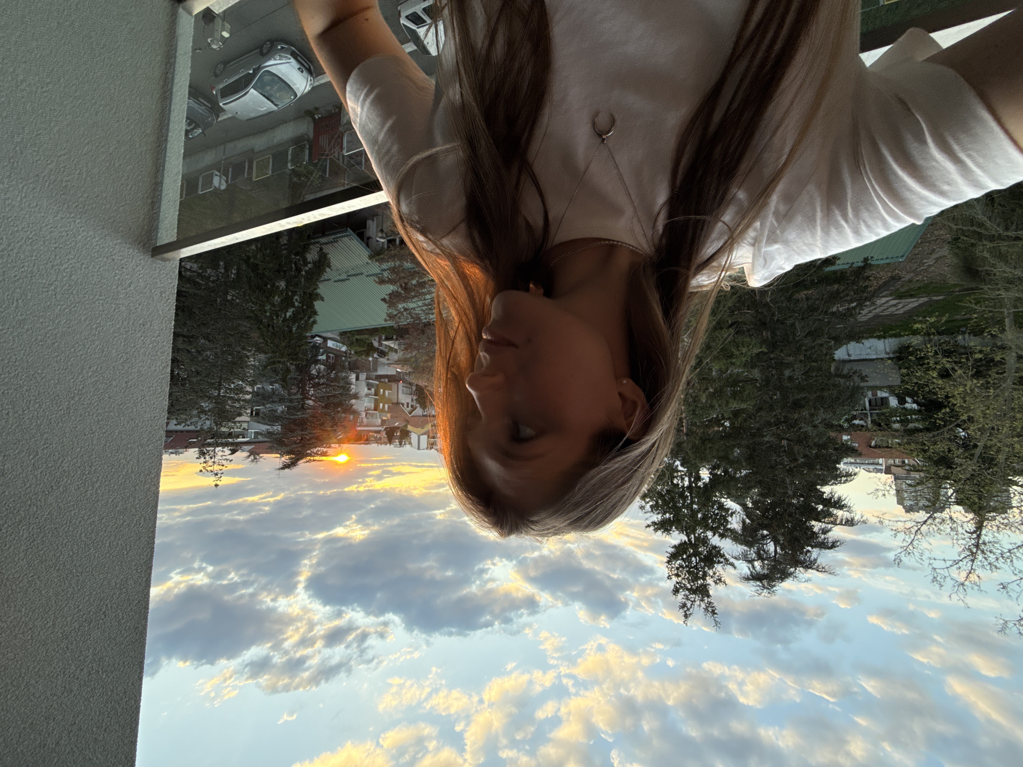 The image, when correctly oriented, depicts a young woman observing a sunset from what appears to be a balcony or window overlooking an urban landscape. The subject, a woman with long, light brown hair, is positioned on the right side of the frame, seen mostly in profile. She is wearing a white short-sleeved top and a necklace with a distinctive crescent moon pendant. Her gaze is directed towards the left, where the sun is setting.
The time of day is clearly golden hour or sunset, with the sun appearing as a bright orange orb low on the horizon. The sky is filled with a beautiful array of clouds, illuminated by the setting sun in shades of white, grey, and brilliant orange-yellow, creating a dramatic and warm atmosphere. Below the horizon, an urban or suburban setting unfolds, featuring various buildings, dense trees, and what appears to be a street with parked cars. A textured grey balcony wall or building facade occupies the foreground on the far left.
The overall scene conveys a sense of peaceful contemplation. The setting, in Vozdovac Urban Municipality, Serbia, is characterized by its blend of built environment and natural elements during a picturesque sunset. There is no visible text in the image.