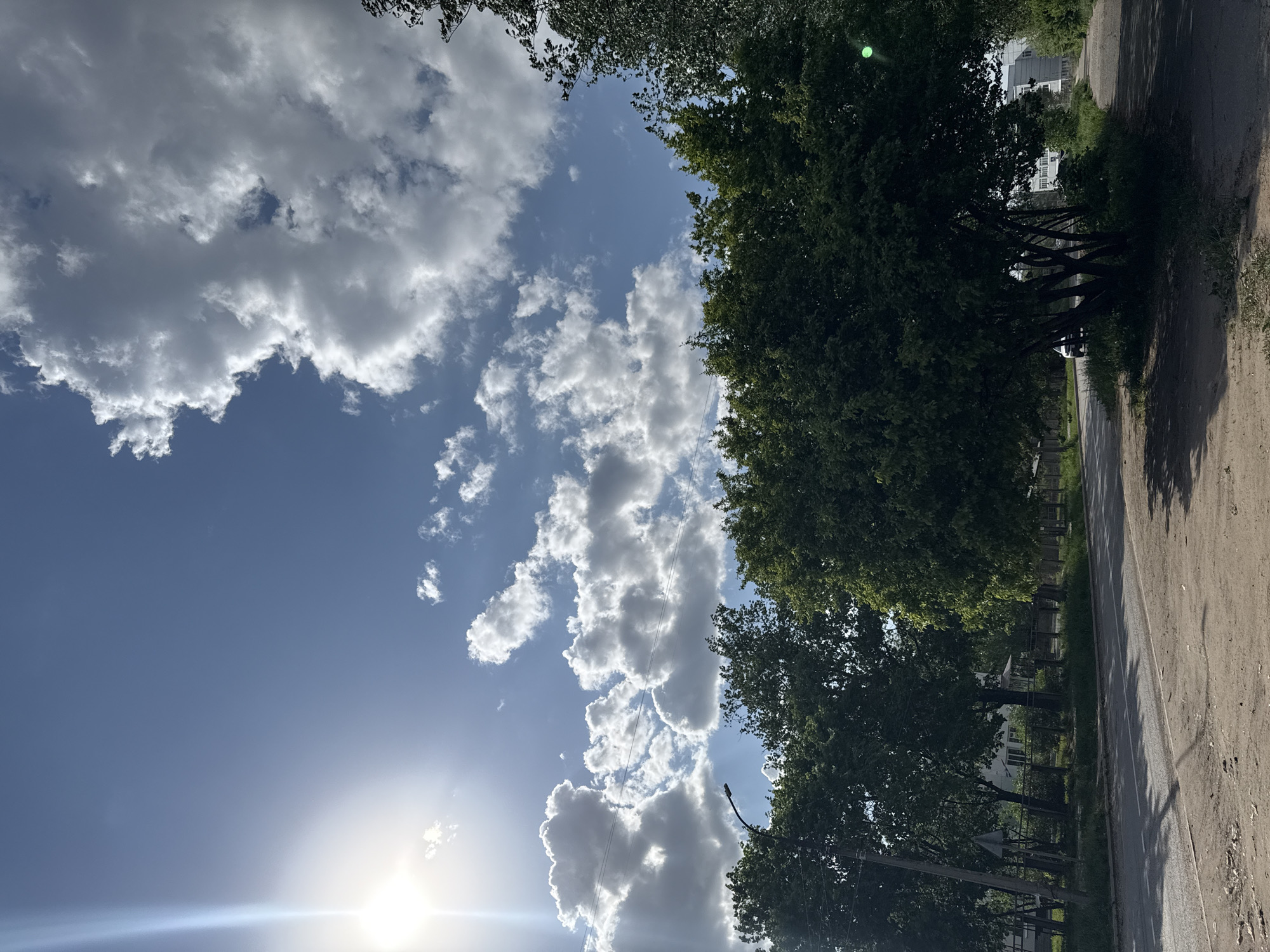 The image shows a view of the sky and a street in Chaikovsky, Russia. The sky is partly cloudy with the sun shining brightly from the lower left corner. To the right is a row of green trees. A road is visible on the right side of the image, with some houses and buildings in the background behind the trees. The image is slightly rotated counter-clockwise.