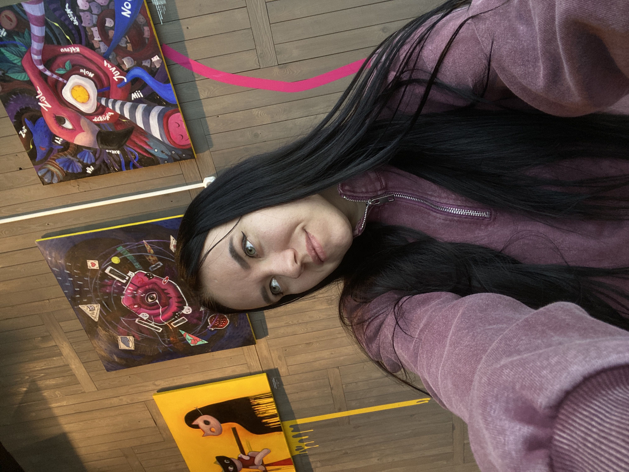 A young woman with long black hair and fair skin is posing for a selfie in what appears to be an art gallery. She is wearing a mauve-colored zip-up sweatshirt. Three paintings are visible on the wooden floor around her. One painting features abstract imagery, including a large red shape with an egg-like element and various written words. Another painting has a dark background with geometric shapes and a stylized rose. The third painting is mostly yellow and shows cartoon-like figures. Pink and yellow tape lines the floor near the paintings. The location is likely the city of Perm, Russia, given the writing in Russian on one of the paintings.