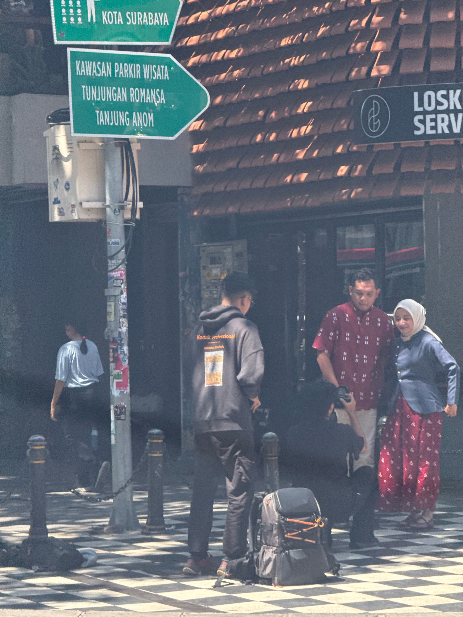 The image shows a street scene in Surabaya, Indonesia, with a focus on a street sign and a group of people. The street sign, in green, reads "KOTA SURABAYA" at the top, and below it "KAWASAN PARKIR WISATA TUNJUNGAN ROMANSA TANJUNG ANOM" pointing to the right. To the right of the sign, a dark sign with a white logo and "LOSK SERV" is visible. In the foreground, a man in a dark hoodie is taking a photo with a camera, and a backpack is placed on the checkered pavement. To his right, a man in a red patterned shirt and a woman in a blue jacket and headscarf are standing. Another person in a light blue shirt is visible further to the left, near a pole covered in stickers. The background shows the tiled roof of a building. The overall scene suggests an outdoor urban environment, possibly a tourist area given the signage.