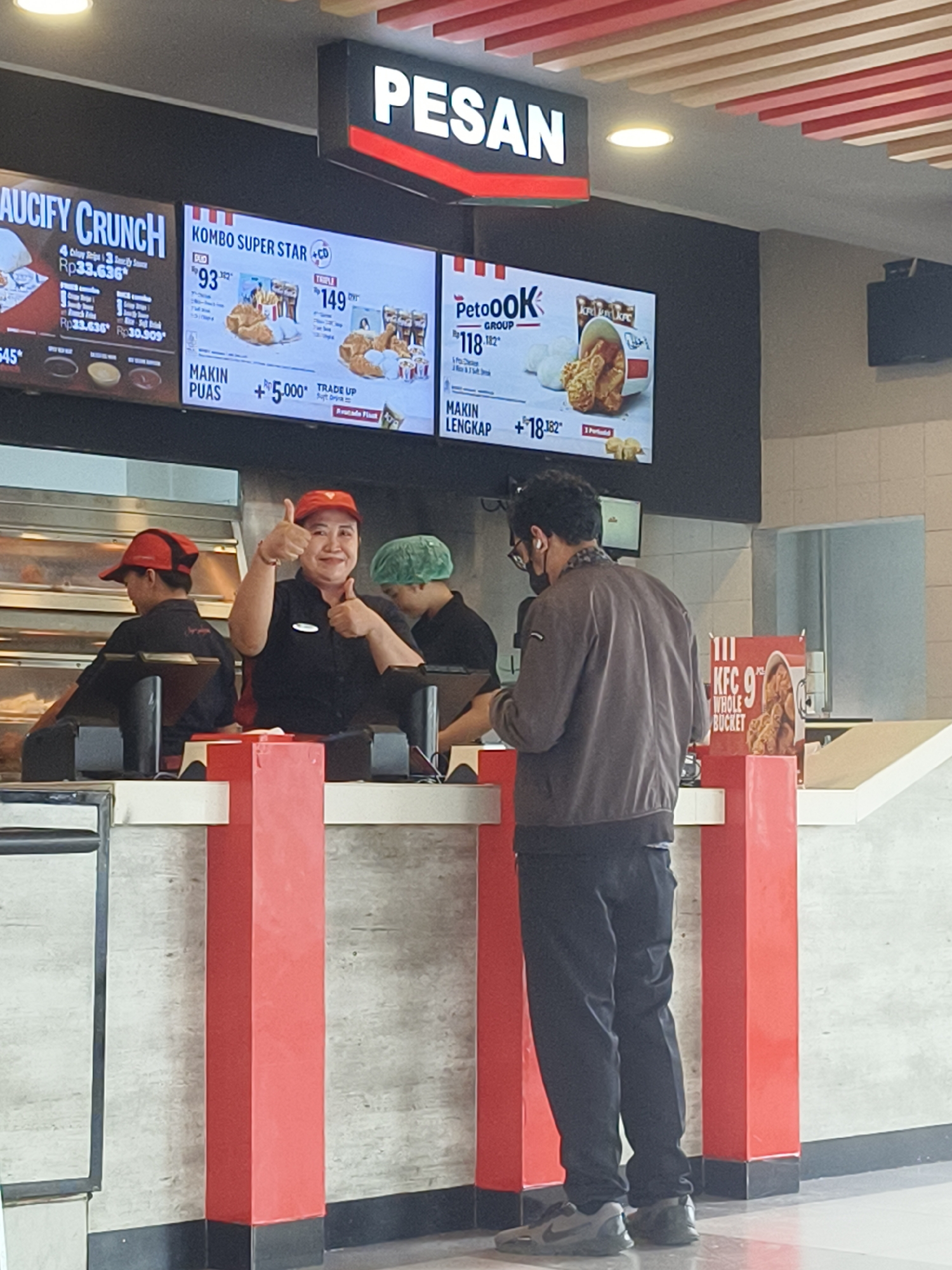 The image depicts the interior of a KFC restaurant in Denpasar, Indonesia, specifically focusing on the ordering counter area. A KFC employee, wearing a red cap and a black uniform, is visible behind the counter, giving a thumbs-up gesture. Two other employees, one in a red cap and the other in a green hairnet, are also present behind the counter. In front of the counter, a male customer, wearing a dark brown jacket and black pants, is standing, facing away from the camera, appearing to be ordering or waiting for his food. Digital menu boards are displayed above the counter, showcasing various KFC meal deals and prices. The restaurant's interior features red and white decor, characteristic of the KFC brand, with red pillars supporting the counter area. The overall scene suggests a typical fast-food restaurant environment with employees serving customers.