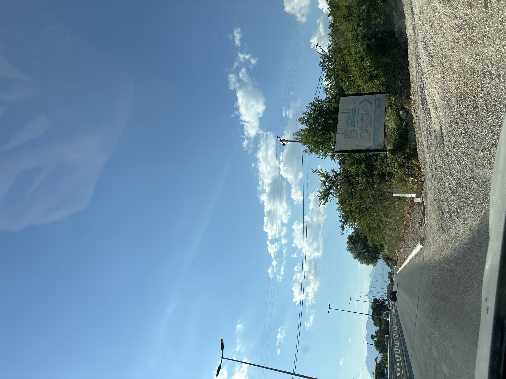 The media file displays a roadside scene under a clear blue sky dotted with white, puffy clouds, suggesting a sunny day. The perspective is from inside a vehicle, looking out towards a road that stretches into the distance, flanked by green trees and shrubs on the left and a gravelly verge on the right. In the distance, mountains are faintly visible, hinting at a rural or semi-rural mountainous region.
Several streetlights line the side of the road, and power lines run overhead. A sign is mounted on a pole to the left of the road, partially obscured by trees. The sign features a logo and text, which appears to read "SMART" and "project>design> (construction)". Below this, a phone number is visible: "+382 67 380 777".
Vehicles are traveling on the road, indicating it is a functional route. The overall impression is one of travel on a bright, possibly warm, day in a scenic area. There are no people clearly visible, but the context suggests a journey or transit. The time of day appears to be late morning or early afternoon, given the bright sunlight. The scene evokes a sense of movement and openness, typical of a road trip.