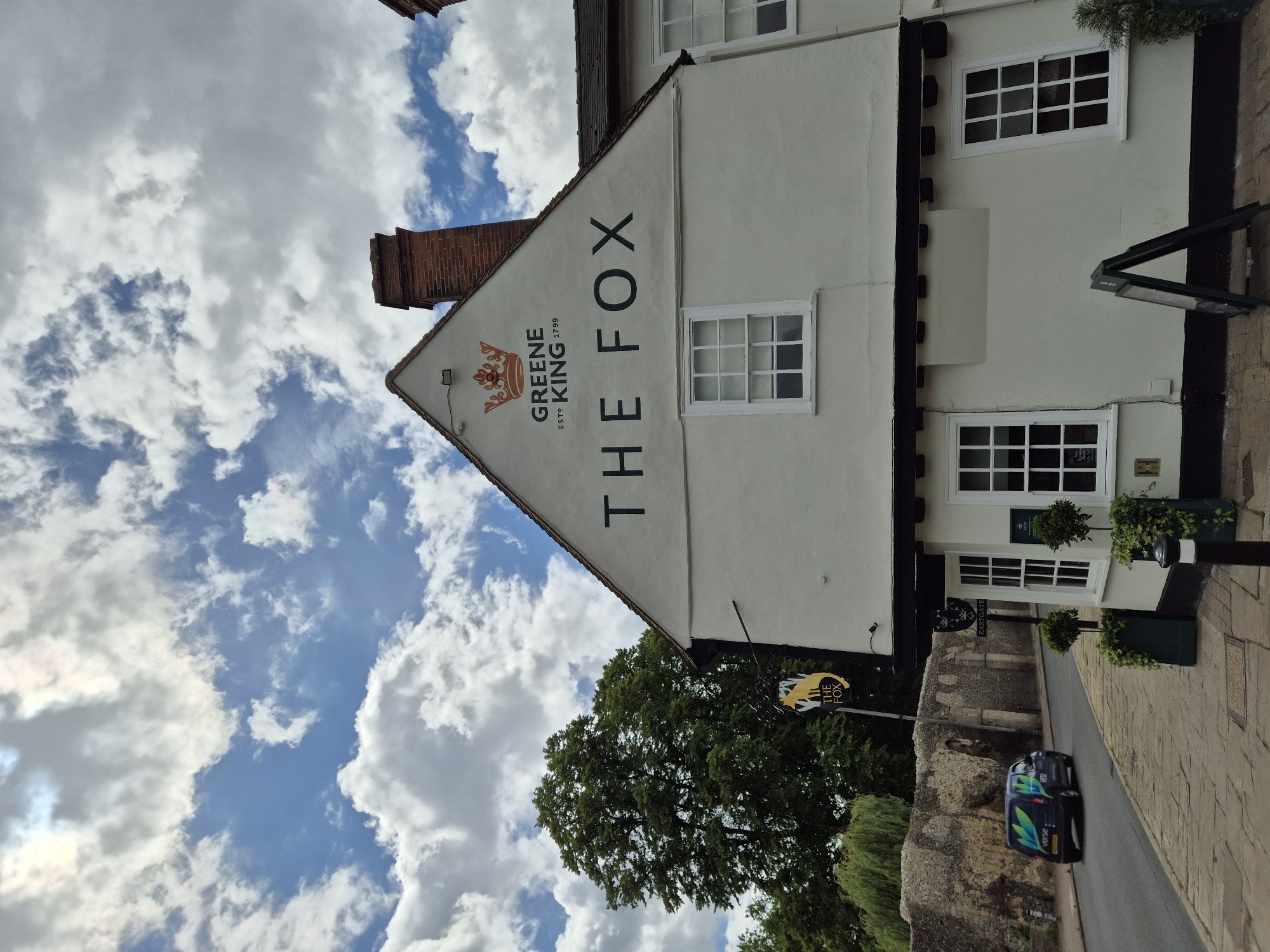 This outdoor image captures the facade of "The Fox" pub, a traditional British establishment, under a partly cloudy sky during the daytime. The building, painted in a light cream or white color, features multiple white-framed windows with small panes and dark trim. The name "THE FOX" is prominently displayed vertically on the side of the building in large black letters, accompanied by the "GREENE KING ESTº 1799" logo, which includes a stylized crown. A brick chimney is visible on the roof.
A hanging sign near the pub's entrance also features "THE FOX" and a stylized fox head in black and yellow. The pub is situated alongside a paved street, where two dark-colored vans are parked. Partial text like "VERSO" and "VEGA" along with green and blue graphics can be seen on the vans. A robust stone wall, suggesting historical architecture, runs parallel to the street opposite the pub. Green trees and foliage are abundant around the area, contributing to a natural setting. Potted plants add decorative touches to the pub's exterior.
The sky is a mix of bright blue and white, fluffy clouds, indicating fair weather. The scene is quiet, with no people visible or specific activities taking place, portraying a calm moment in what appears to be a historic village or town setting in West Suffolk, United Kingdom.