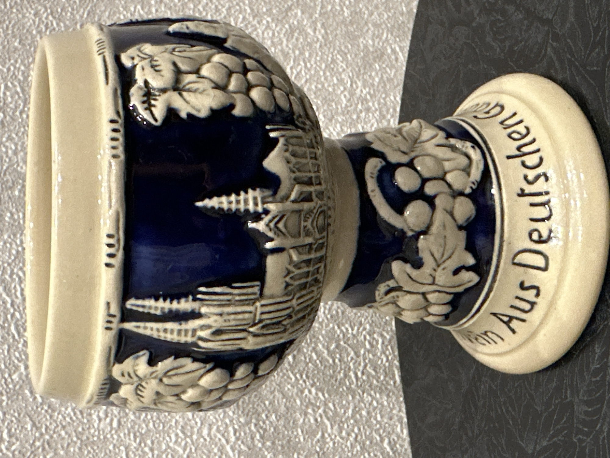 A close-up, eye-level shot shows a decorative ceramic stein with a blue body and a beige rim and base. The stein features embossed images of grapevines and what appears to be a cathedral or castle. Along the base, in black lettering, the words "in Aus Deutschen" are visible, suggesting a German origin or theme. The stein rests on a dark, textured surface, possibly a table. The background is a textured, light-colored wall. There are no people present in the image, and the object is the sole focus. The image is well-lit, with natural light contributing to the overall clarity. The location in the city of Energetik, Russia, is not ascertainable from the visual information provided in the image.