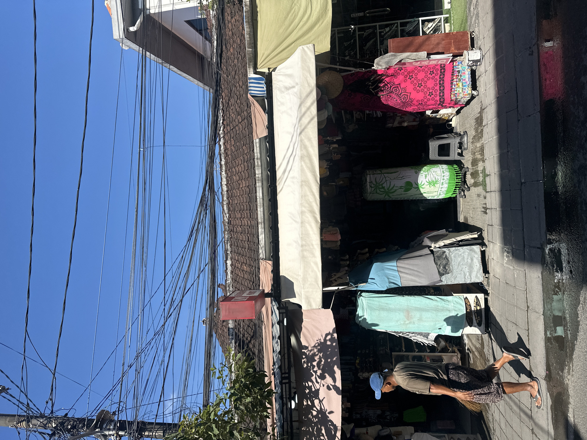 In the city of North Kuta, Indonesia, a man wearing a brown t-shirt, patterned shorts, a blue baseball cap, and flip-flops walks along a tiled sidewalk, sweeping the ground with a broom. He is moving from left to right. The sidewalk is wet in some areas, reflecting the bright sunlight. To his right, a street-side shop displays various items. The shop has a white awning providing shade, and beyond it, clothing and textiles are visible, including a bright pink garment with a black floral pattern and a green cylindrical display with a white palm tree design. The shop is covered by a tiled roof, and above the shop, a complex network of electrical wires crisscrosses against a clear blue sky. A red sign with white lettering hangs above the shop. The building's facade is partially visible, with modern architectural elements contrasting with the traditional tiled roof. The scene suggests a typical day in a commercial area, with an emphasis on daily life and commerce.
