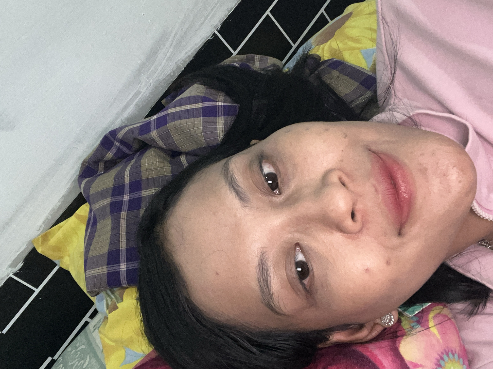 The image is a close-up, high-angle selfie of a woman lying on a bed. She has dark hair, brown eyes, and is wearing a pink shirt and a diamond stud earring. The background shows a yellow and purple plaid blanket, and a wall with black and white tiles. The scene is indoors and appears to be in a bedroom. The woman is looking directly at the camera with a slight smile. The image does not provide any information about the city of Yogyakarta, Indonesia.
