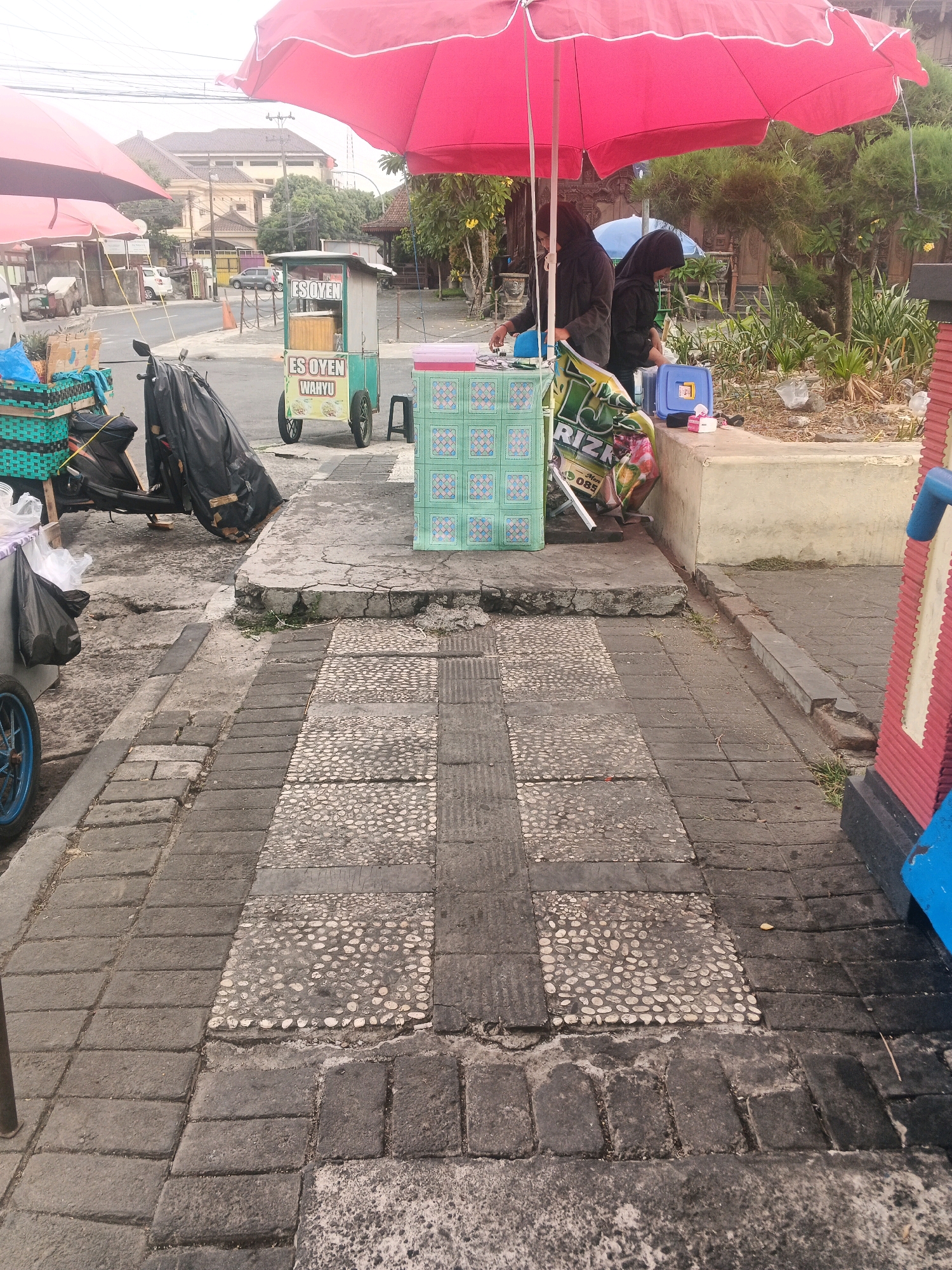 The image depicts a street scene in Ungaran, Indonesia, with a focus on a small food stall. A red umbrella shades two individuals, likely vendors, who are attending to the stall. The stall itself is a cart with the text "ES OYEN WAHYU" displayed, indicating it sells a local beverage. Various items are visible around the stall, including plastic crates, a motorcycle covered with a black tarp, and a blue cooler. The ground is paved with a combination of large rectangular stones and smaller pebble-like stones, with some areas showing wear and tear. In the background, buildings and trees suggest an urban environment. The overall impression is a typical roadside vendor operation in a town or city setting.