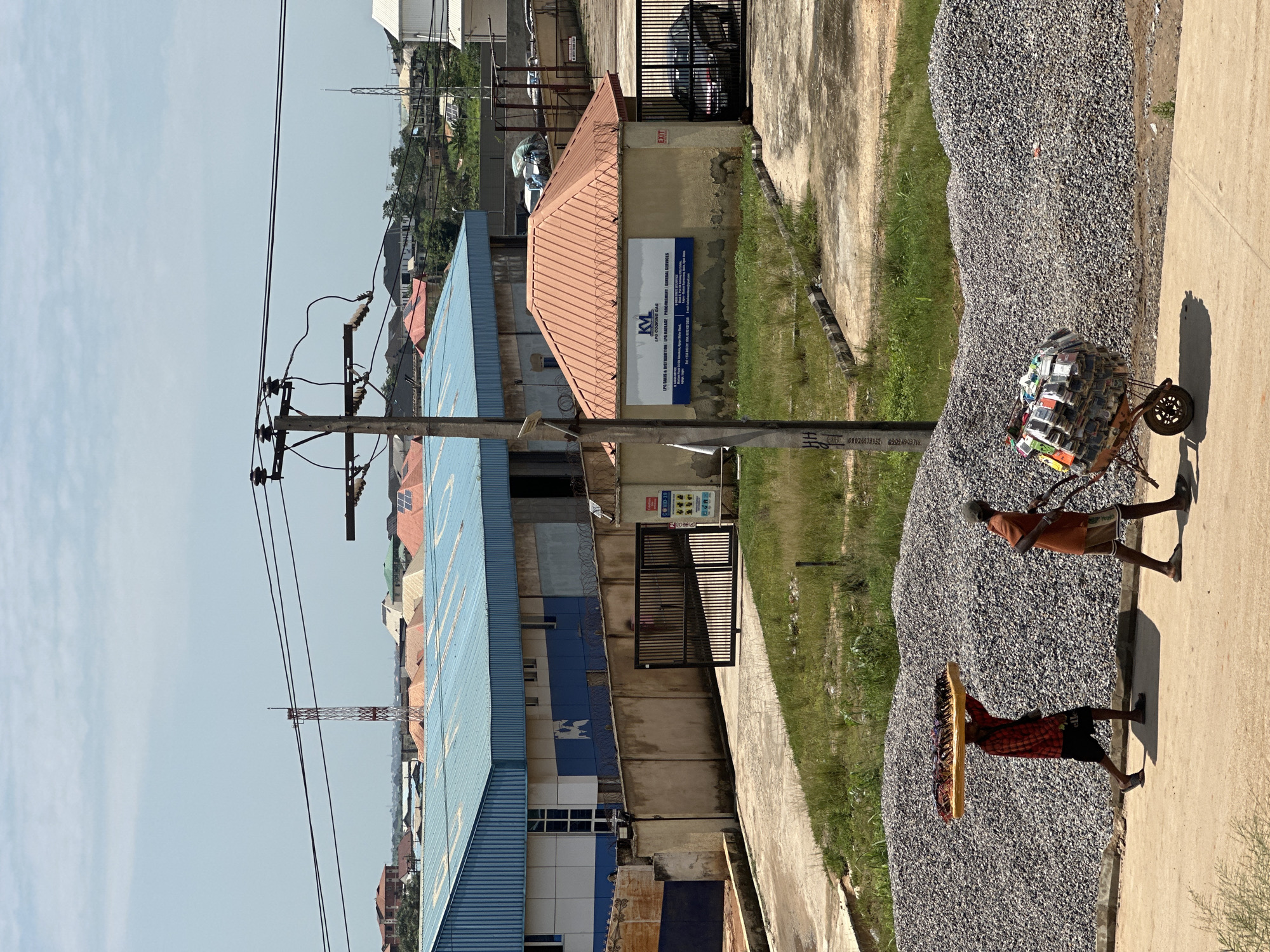 In Magboro, Nigeria, two men are seen working near a construction site. The man on the left carries a large, flat object, possibly a mattress, balanced on his head. He wears a red and black checkered shirt and black shorts. To his right, another man pushes a wheelbarrow laden with a stack of items, which appear to be phone cases or small electronics. He is dressed in an orange t-shirt and beige shorts. They are walking on a concrete path in front of two large mounds of gray gravel. In the background, a long, industrial building with a blue roof and a smaller building with an orange roof are visible. Power lines stretch across the sky above the structures, and a telecommunications tower can be seen in the distance. The environment appears to be an urban or semi-urban area under a bright, cloudy sky.