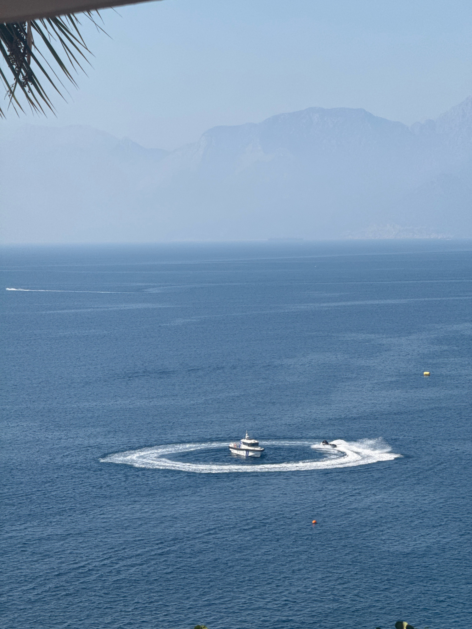 A high-angle, medium shot captures a tranquil expanse of deep blue sea under a pale blue sky. In the foreground, the fronds of a palm tree are visible in the upper left corner, casting a shadow. The middle ground is dominated by the ocean, with a white motorboat making a sharp turn, leaving a swirling wake behind it. A jet ski follows closely, also creating a significant wake. A small orange buoy is present in the water. In the distance, hazy blue mountains form the horizon line. The scene appears to be the coast of Antalya, Turkey, likely a popular spot for watersports.