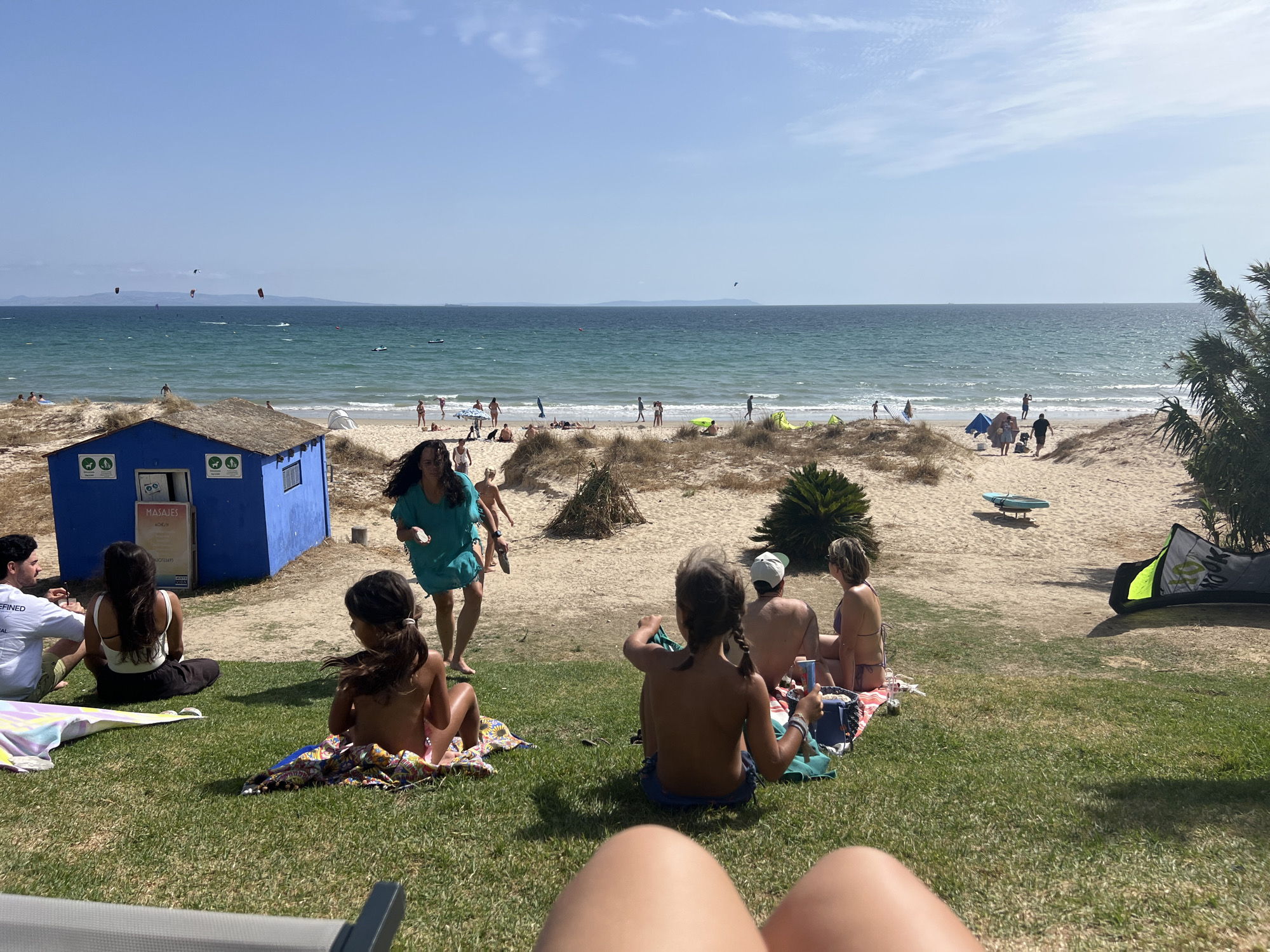 The image depicts a sunny day at the beach, likely in Tarifa, Spain, given the presence of kitesurfers visible in the distance and the coastal landscape. In the foreground, several people are relaxing on the grass, some sitting on blankets. A young girl with braided hair is in the center, facing away from the camera. To her left, another child is also seated, facing away. To the left of them, a man and a woman are seated, the man wearing a white t-shirt. In the mid-ground, a blue beach hut with a thatched roof is visible. A woman in a teal dress is standing near the hut, appearing to interact with someone out of frame. The background features a sandy beach with scattered vegetation, more people, and the calm blue ocean. Kitesurfers are actively engaged in the water further out. The overall atmosphere is relaxed and recreational, characteristic of a beach destination.