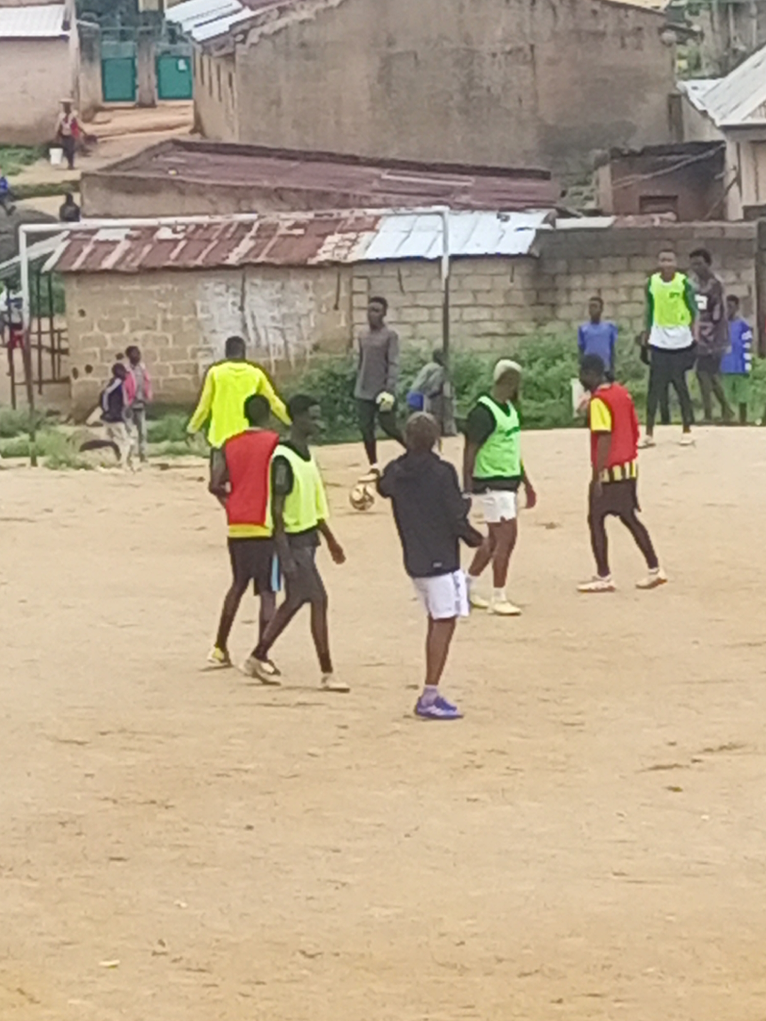 A group of young men are playing soccer on a dirt field in Jos, Nigeria. The field is surrounded by low-rise buildings with corrugated metal roofs. The players are wearing casual clothing, including t-shirts, shorts, and soccer cleats. One player is wearing a bright yellow shirt, another a red vest, and several are wearing green vests. The sky is overcast, and the lighting suggests it is daytime. The overall scene depicts a casual pickup soccer game in an urban environment.