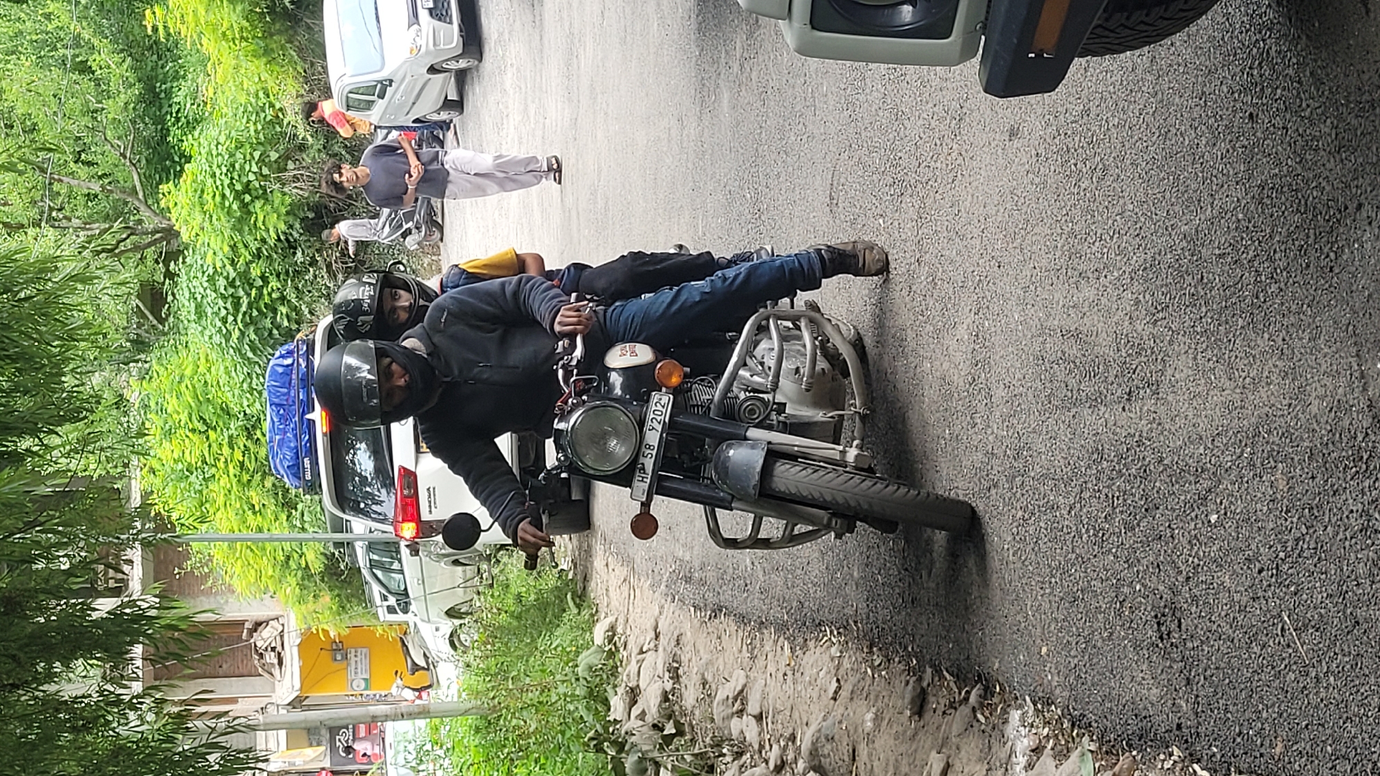 In Vashisht, India, on a paved road, a man wearing a black helmet and a dark jacket rides a dark-colored Royal Enfield motorcycle. A younger person, also wearing a black helmet, sits behind him. The motorcycle has a license plate reading "HP 58 Y 202". In the background, a white SUV with a blue carrier on top is parked, and other vehicles and people are visible on the road, along with dense green foliage. The scene suggests a moment captured on a street in a mountainous or hilly region.