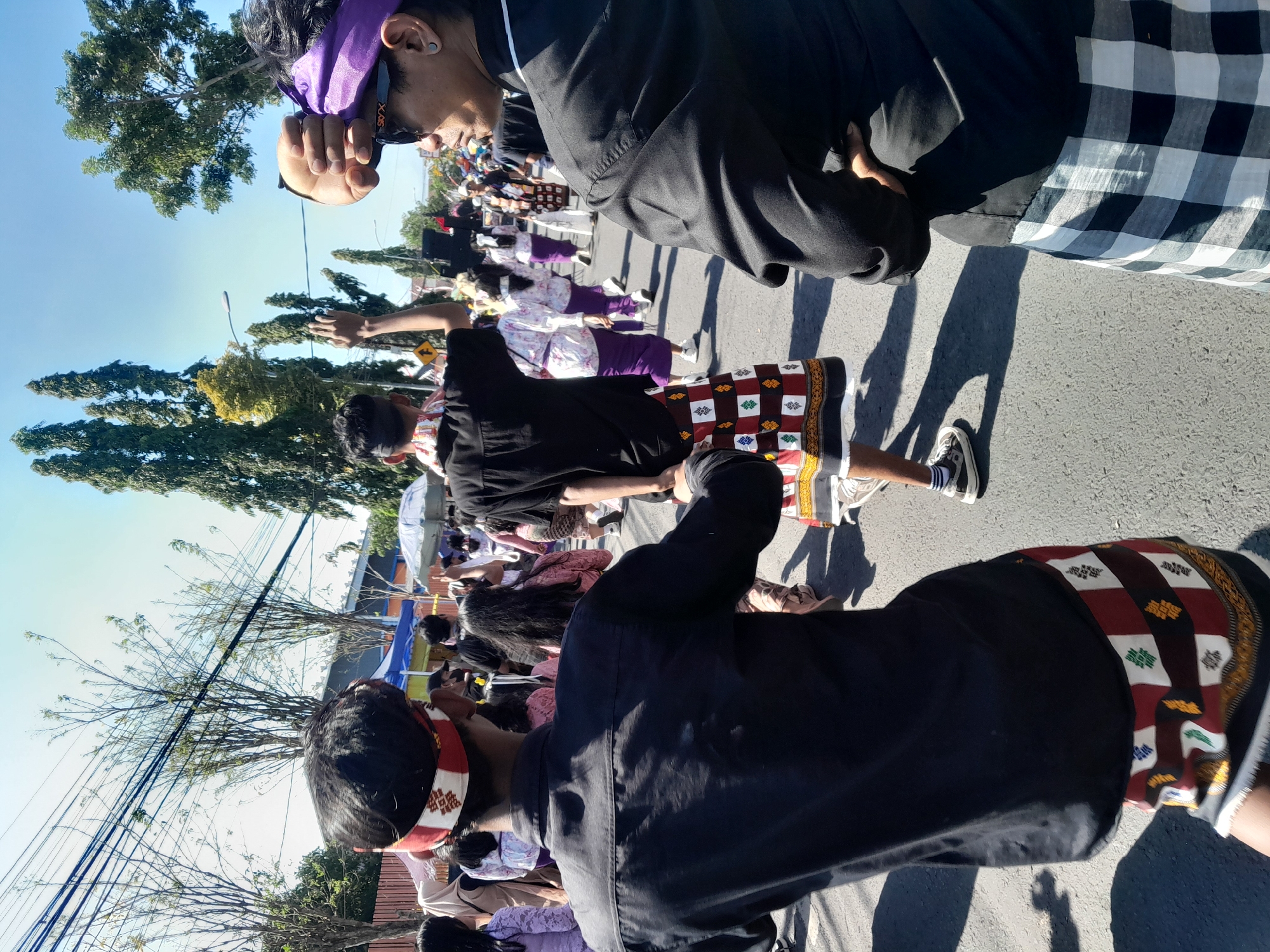 In the city of Gentengwetan, Indonesia, under a bright sky, a procession moves down a paved street. The foreground features a person from behind, wearing a black shirt and a distinctive skirt with a red and white geometric pattern. To their right, another person, also seen from the side, wears a black shirt, sunglasses, and a purple headband. Their hands are clasped in front of them. The middle ground reveals a group of individuals, some wearing similar patterned skirts in purple and white. They appear to be part of the procession, walking or dancing. In the background, tall, slender trees line the street, and overhead wires crisscross the sky. Distant buildings and tents suggest an event or festival taking place. The sun casts long shadows on the asphalt, indicating it is likely daytime.