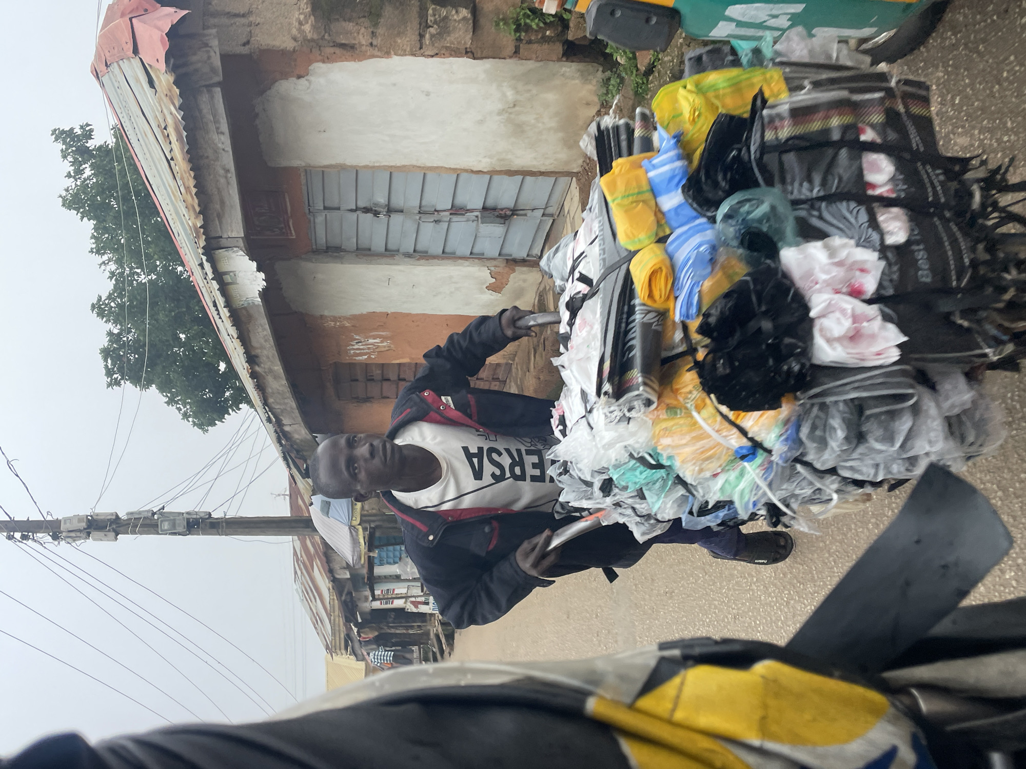In Jos, Nigeria, on a street with a cloudy sky, a young man is pushing a cart laden with various plastic bags and bundles of goods. He is wearing a black jacket with red trim over a white t-shirt that reads "VERSA" in black letters. The cart is full of an assortment of colorful plastic bags, some of which are yellow, blue, and white striped, and black trash bags. Behind him, a portion of a vehicle, possibly a motorcycle, with yellow and black coloring is visible in the foreground. The street is lined with buildings constructed with adobe and corrugated metal roofs. Power lines are strung across the scene, supported by a utility pole with multiple meters attached. Some of the buildings appear to be small shops or residences. The overall impression is a bustling street scene in a developing urban environment.