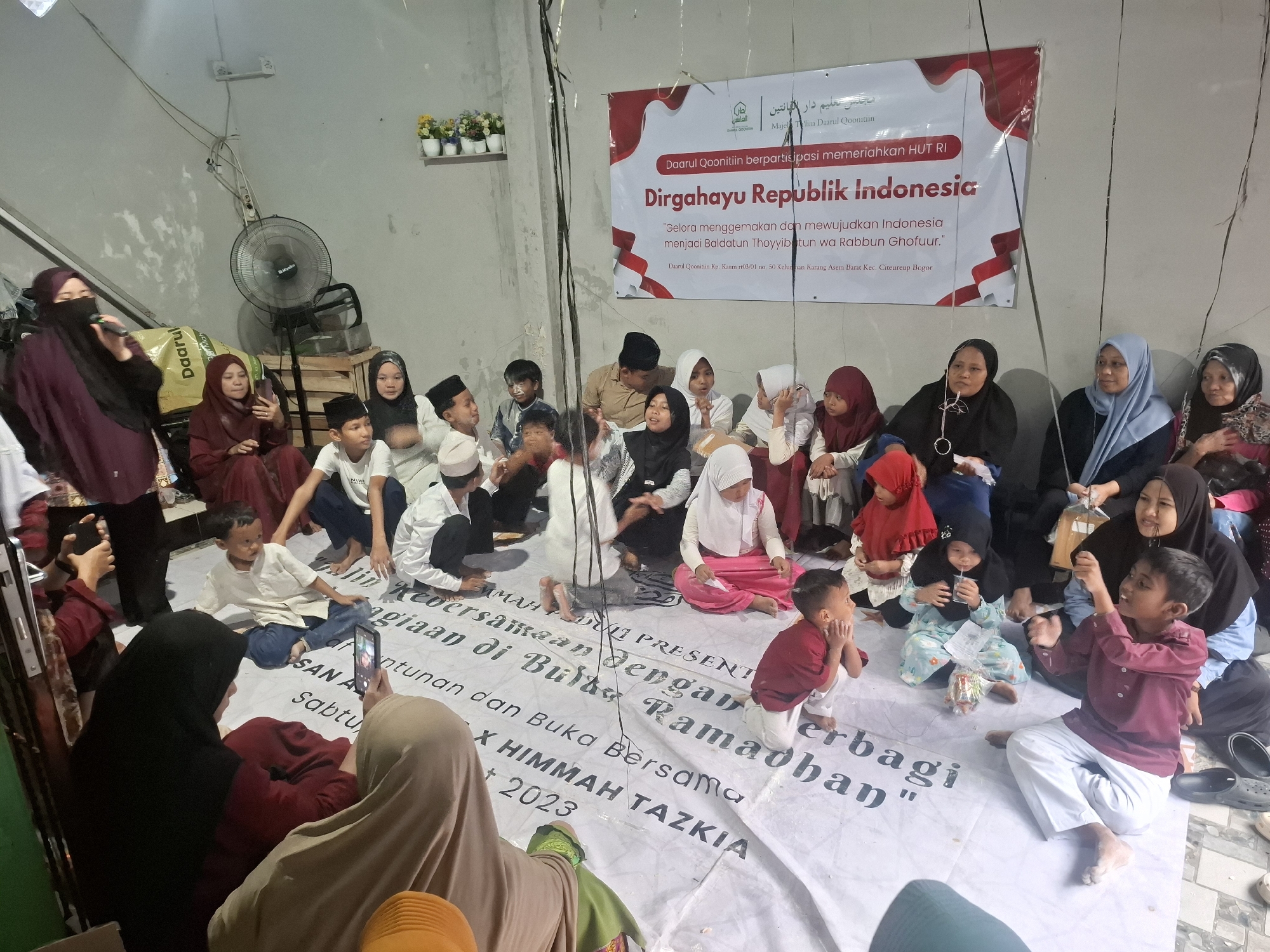 This is a wide shot of a group of people gathered on the floor, likely for an event. A large banner hangs on the wall behind them, which appears to be a celebration of Indonesian Independence Day. Several children are in the foreground, some are sitting on the floor, and others are being held by adults. A woman in the upper left corner is holding a microphone, and it appears she might be speaking. The room is sparsely decorated, with some plants on a shelf and a fan in the background. The overall impression is of a community gathering, possibly a religious or cultural event, taking place in a simple indoor setting. The banner mentions "Dirgahayu Republik Indonesia" and "Darul Qooniitiin," suggesting a connection to a religious institution named Darul Qooniitiin and the Indonesian national day. The location is identified as Babakan Madang, Indonesia.