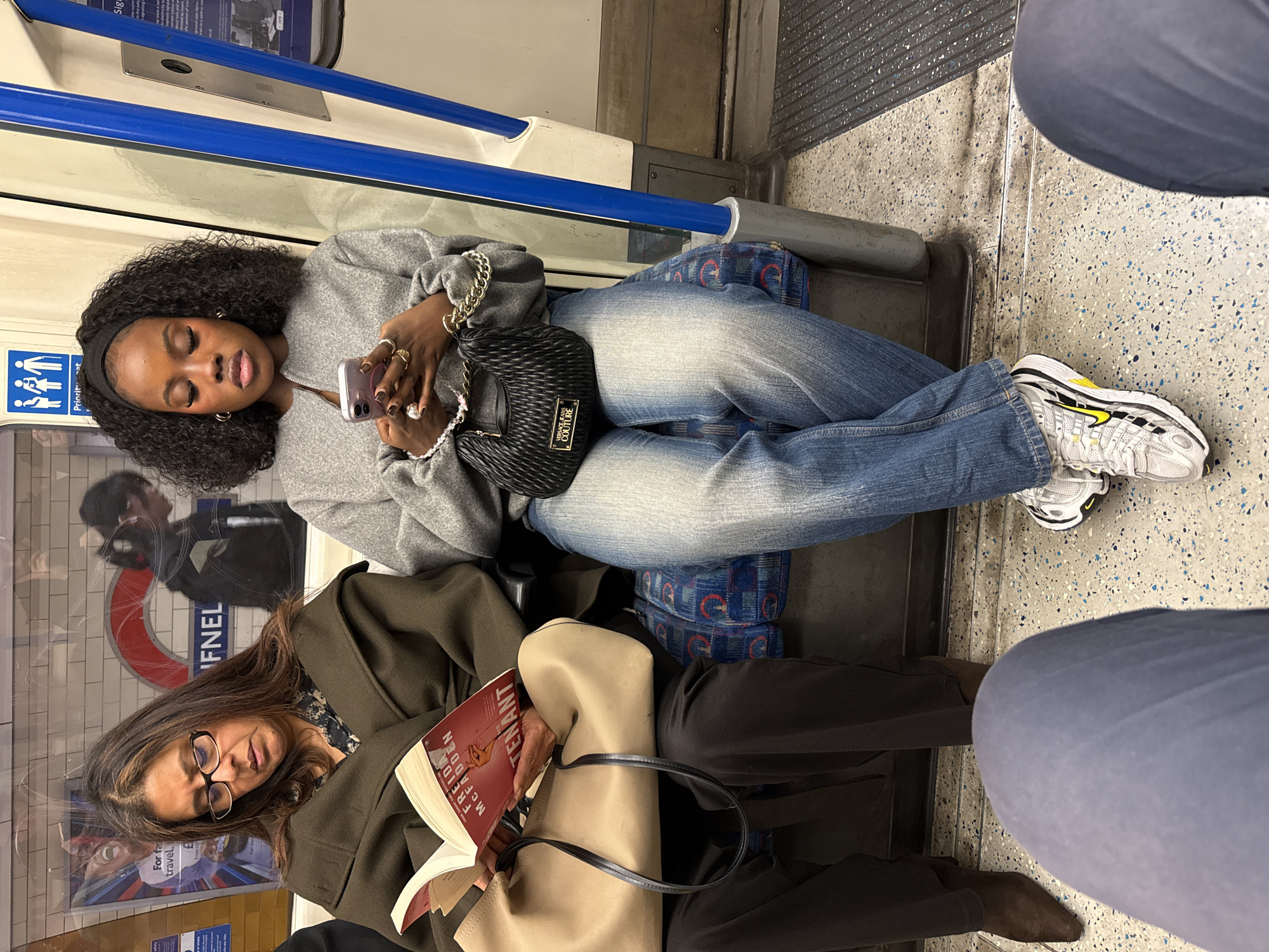 The image depicts two individuals seated on a London Underground train. The person at the top of the image is a young Black woman using a mobile phone. She wears a grey sweatshirt, light blue jeans, white sneakers with a yellow stripe, and carries a Versace Jeans Couture handbag. She has multiple rings on her fingers and a thick, silver-toned bracelet. The person at the bottom of the image is an older woman reading a book titled "The Tenant" by Freida McFadden. She wears glasses, a brown coat, black trousers, and is carrying a large beige tote bag. The London Underground roundel with "TUFNELL" is visible in the reflection of the window. The train seats have a blue and red patterned upholstery. A third, unidentified person partially appears in the right of the image.