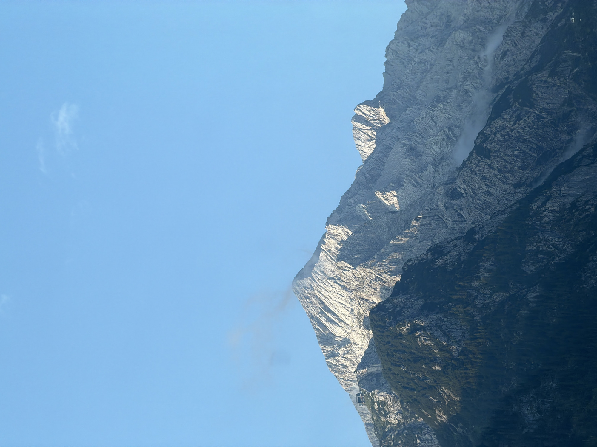 The image shows a majestic mountain peak in Grainau, Germany, under a clear blue sky with a few wisps of clouds. The mountain is characterized by rugged, rocky slopes, with the upper portions illuminated by sunlight, highlighting the textures of the rock formations. The lower slopes are covered in dark green coniferous trees. A small building, possibly a hut or a lodge, is visible on a ledge partway up the mountain. The overall scene conveys a sense of natural grandeur and isolation.