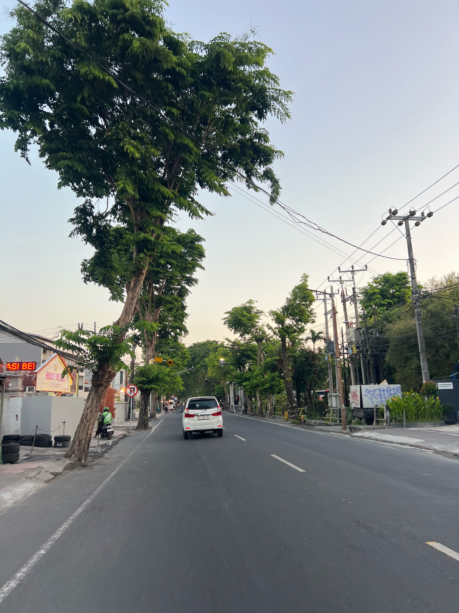 A street scene in Kuta, Indonesia, captures a road lined with trees and utility poles. A white van is driving away from the camera on the asphalt road, which has a white dashed line marking the lane. To the left of the road, a sidewalk is visible with some stacked tires and a building with neon signs, one of which reads "ASI BEI". A person on a green motorcycle with a green bag on their back is stopped near the sidewalk. Utility poles with numerous wires are situated along the right side of the road, interspersed with more trees and foliage. The sky is a pale blue with hints of orange, suggesting it might be early morning or late afternoon.