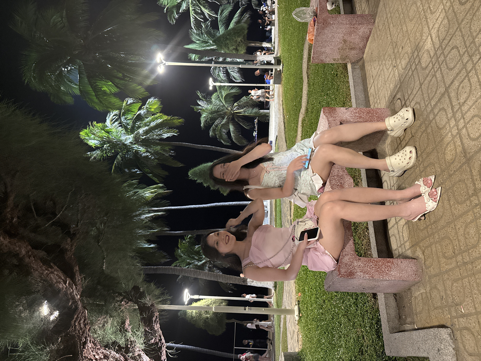 Two young women sit side by side on a public bench in Nha Trang Ward, Vietnam, at night. The woman on the left wears a light pink dress, holds a smartphone, and smiles at the camera. The woman on the right wears a light blue patterned dress and covers her face with her hand. They are sitting on a stone bench with a green patch of grass behind them. The ground is paved with patterned tiles. Palm trees and streetlights are visible in the background, and other people are walking along the pathway behind the bench.