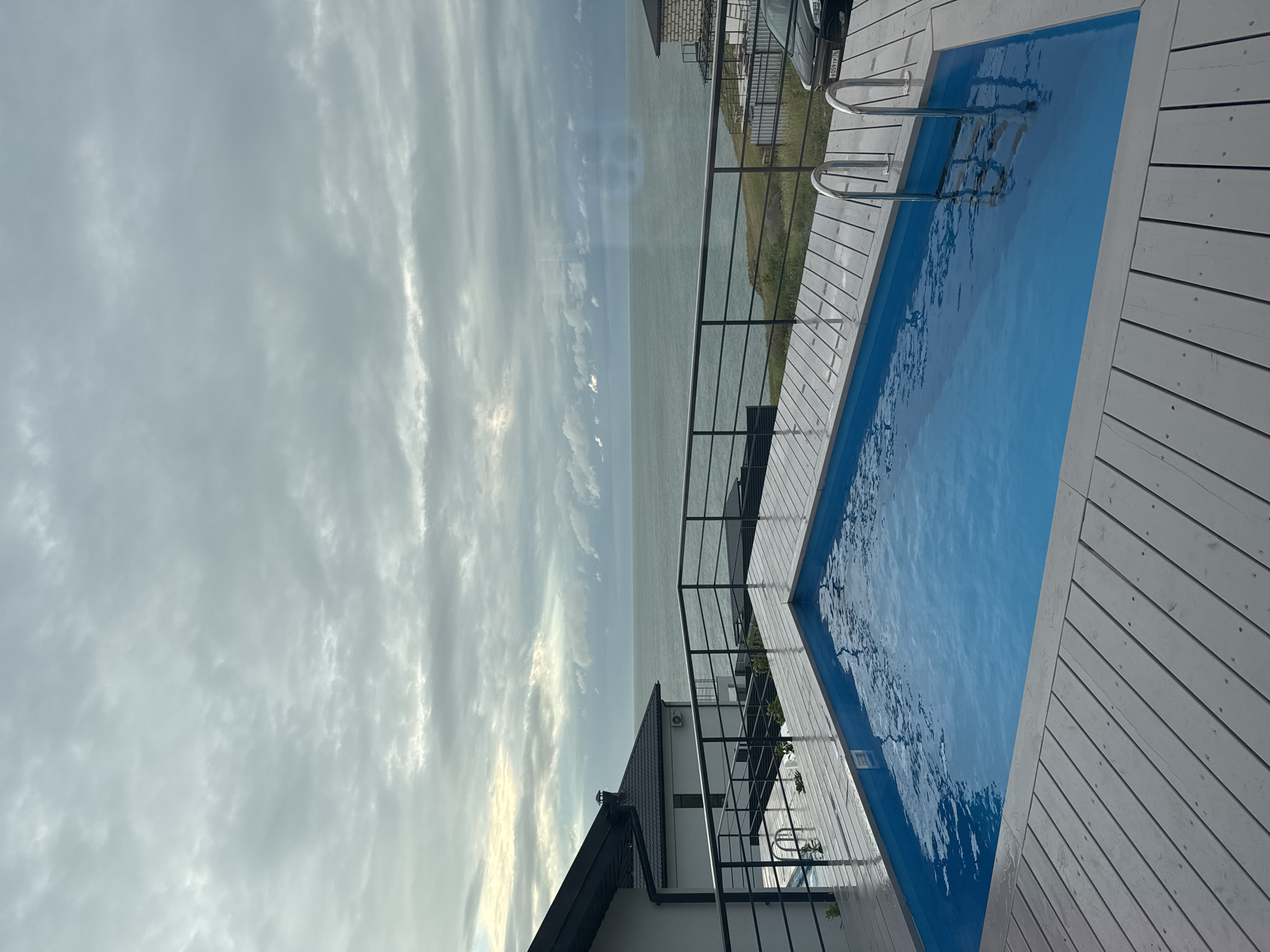 The image depicts a rectangular swimming pool located on a white wooden deck next to a body of water under a cloudy sky. A black metal railing surrounds the deck and the pool has a metal ladder. A white house with a dark gray roof is visible in the background. Based on the available information, the location is Мысовское сельское поселение, Ukraine.
