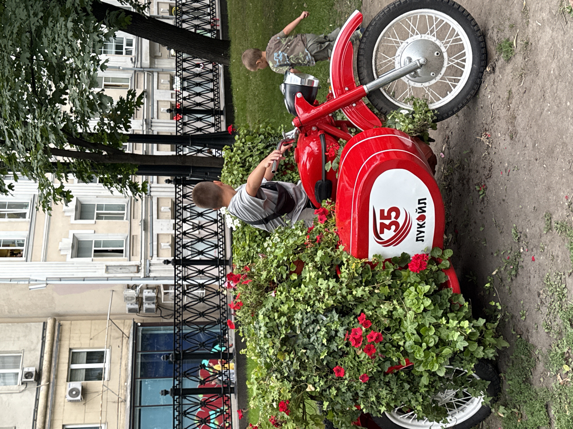 In Moscow, Russia, two young boys are the subjects of this photograph. The boy on the left, facing away from the camera, is positioned behind the handlebars of a vintage red motorcycle with a sidecar. He is wearing a light-colored t-shirt and dark pants, with a small brown bag slung across his chest. The motorcycle is adorned with lush green foliage and vibrant red flowers, partially obscuring the sidecar. A white oval logo with the number "35" and Cyrillic text "ЛУКОЙЛ" is visible on the sidecar.
To the right, a second boy, facing away from the camera and towards the motorcycle, is wearing a camouflage t-shirt and green pants. He appears to be walking or running in the direction of the motorcycle.
The scene is set outdoors, with a paved area in the foreground and a grassy area behind the motorcycle. In the background, a black metal fence separates the immediate area from a sidewalk and a building. The building, with its light-colored facade, features multiple windows and air conditioning units. Trees with green foliage are also present, contributing to the urban park-like atmosphere. The overall lighting suggests it is daytime, possibly overcast.