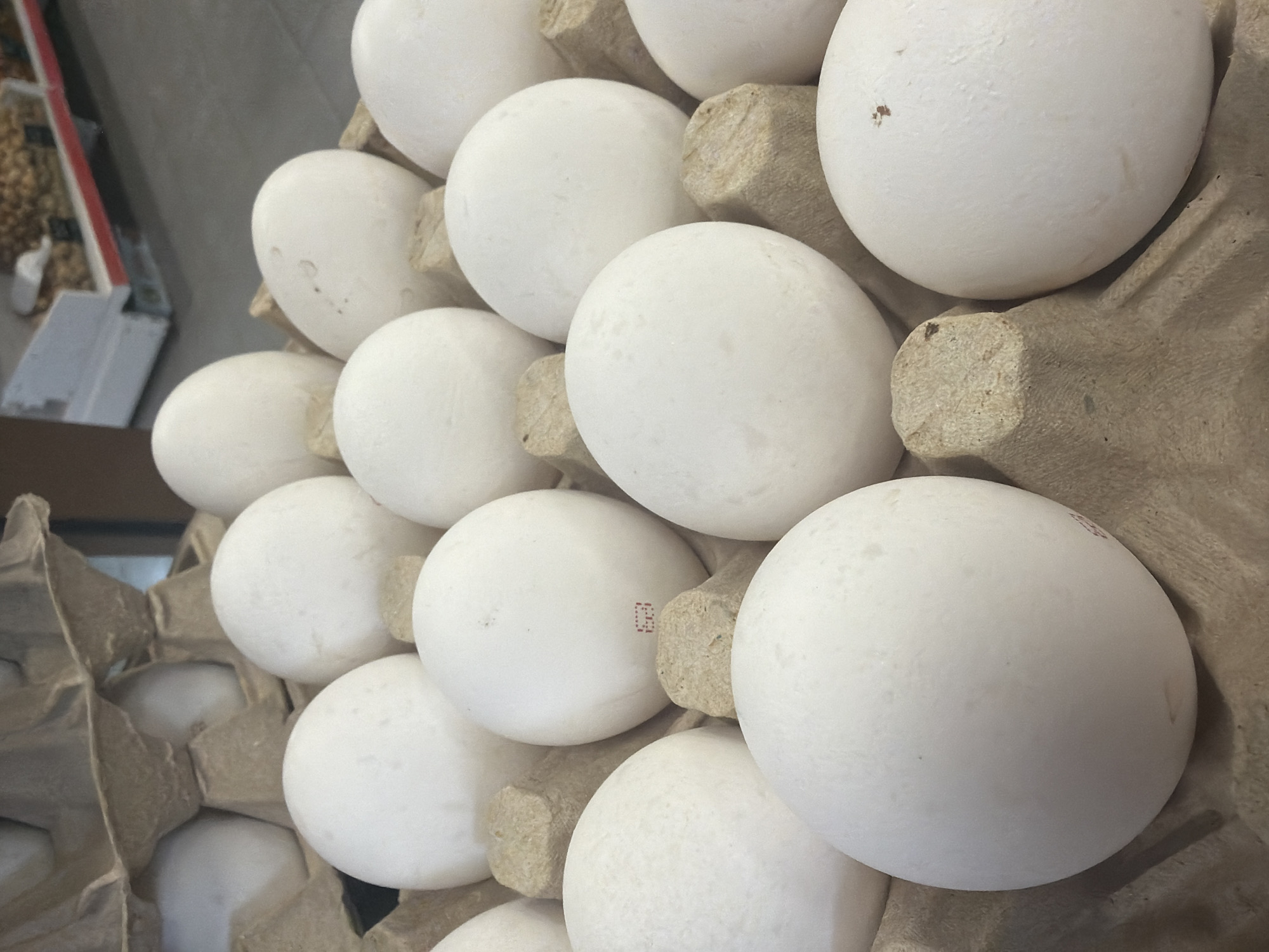 The image shows a tray of white eggs. The eggs are arranged in a cardboard carton. The carton is brown and has a rough texture. The eggs are white and smooth. The image is taken from a close-up perspective. The scene is indoors, possibly in a store or a refrigerator. The eggs are likely for sale or for consumption. The location in the city of Krasnoyarsk, Russia is unknown.