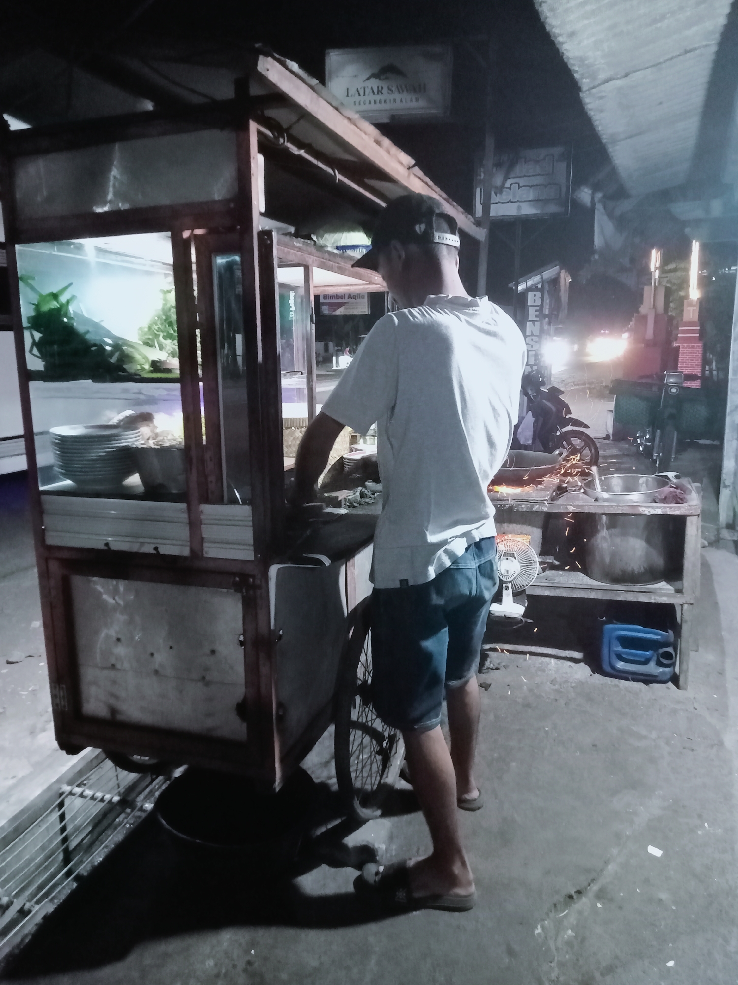 The image shows a food cart at night in Prajeksari, Indonesia. A man wearing a light-colored t-shirt, denim shorts, and a baseball cap stands behind the cart, appearing to be preparing food. Sparks are visible near a metal basin on the cart, suggesting cooking or grinding is taking place. The cart has a glass display case with what appears to be food inside, and a stack of plates. In the background, there are signs, a motorcycle, and building lights. The ground is paved, and there is a blue plastic container near the cart.