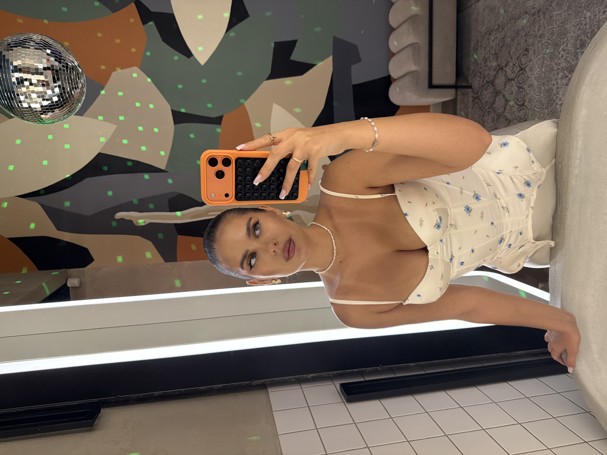 A woman with dark hair pulled back in a ponytail, wearing a white floral corset-style top and a pearl necklace, takes a mirror selfie in a dimly lit room. She holds an orange-cased smartphone in her right hand. Her left hand rests on the edge of a white bathtub. In the background, a large, abstract mural with muted colors and green speckles adorns the wall. A disco ball hangs from the ceiling, reflecting light. The room appears to be a bathroom or dressing area with tiled walls and modern lighting fixtures. The overall ambiance is chic and possibly suggests a pre-event preparation scene. There are no elements in the image that can place it in Krasnoyarsk, Russia.