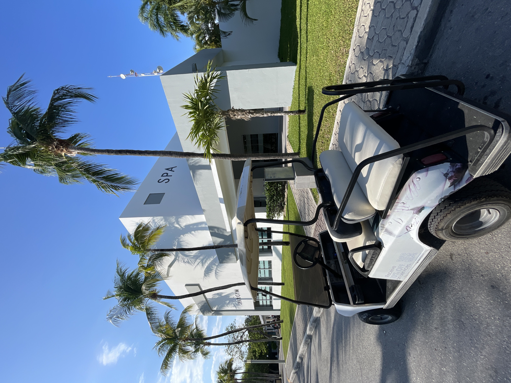 The image depicts a white golf cart parked on an asphalt road in Playa del Carmen, Mexico. The cart has white seats and black trim, and some type of advertisement, either hand-painted or applied as a decal, is visible on the exterior. Behind the golf cart is a modern, white building with palm trees in front. The building has the word "SPA" and "GYM" imprinted on the exterior. The sky is blue, and the lighting suggests a sunny day. The scene appears to be a resort or upscale area.